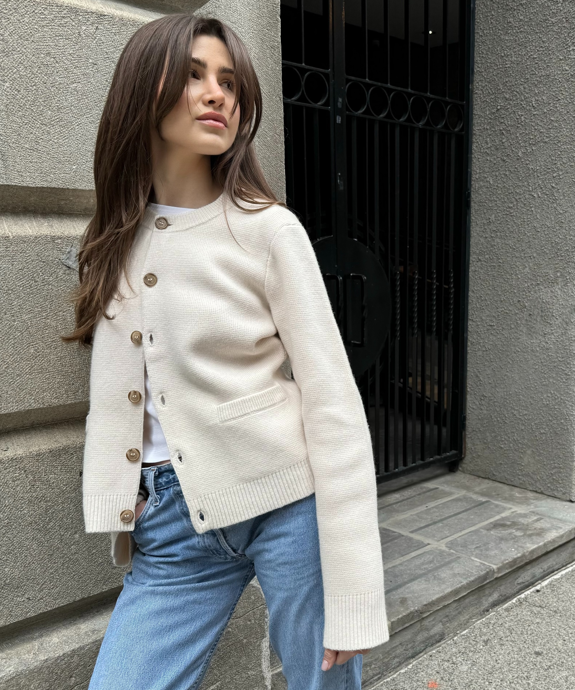 anna porte wearing an ivory cooper cardigan with a white tee and blue jeans leaning against a stone wall