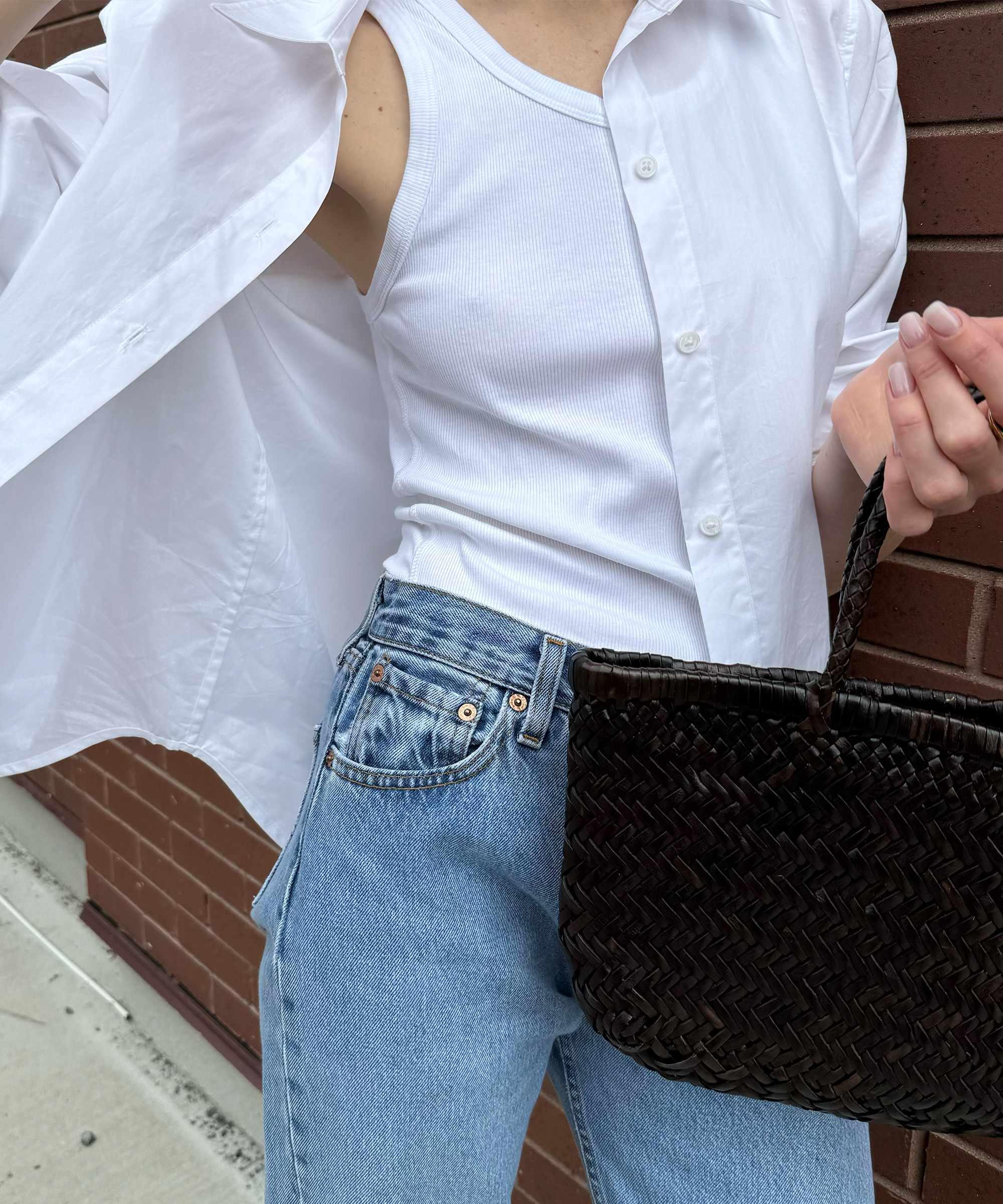 A person wearing a sleeveless white top, an open white button-down shirt, and blue jeans holds a woven dark brown handbag from anna porte while standing near a brick wall. Their face is not visible.