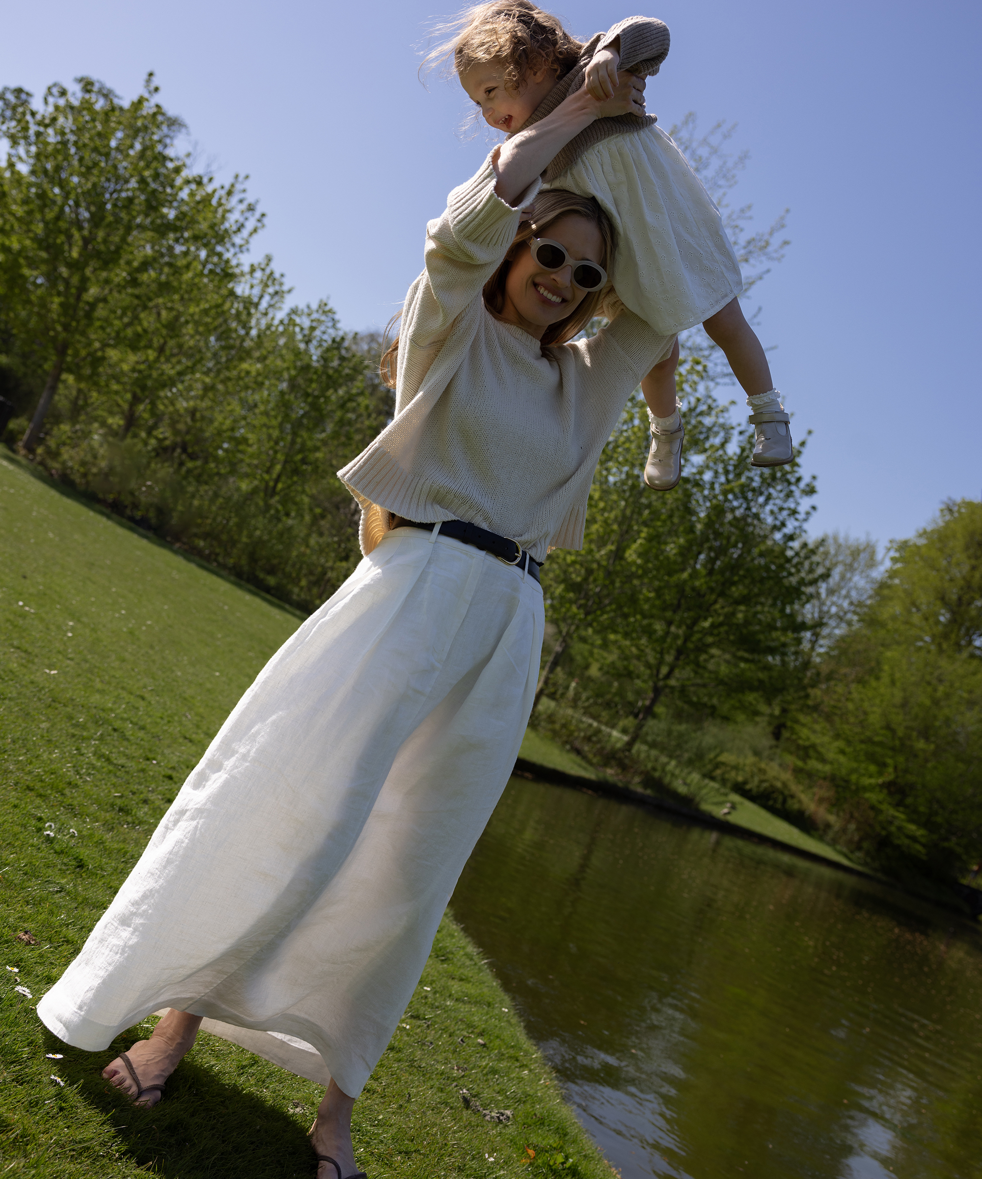 cecilie in a white linen skirt and cream sweater standing in the grass holding up her daughter