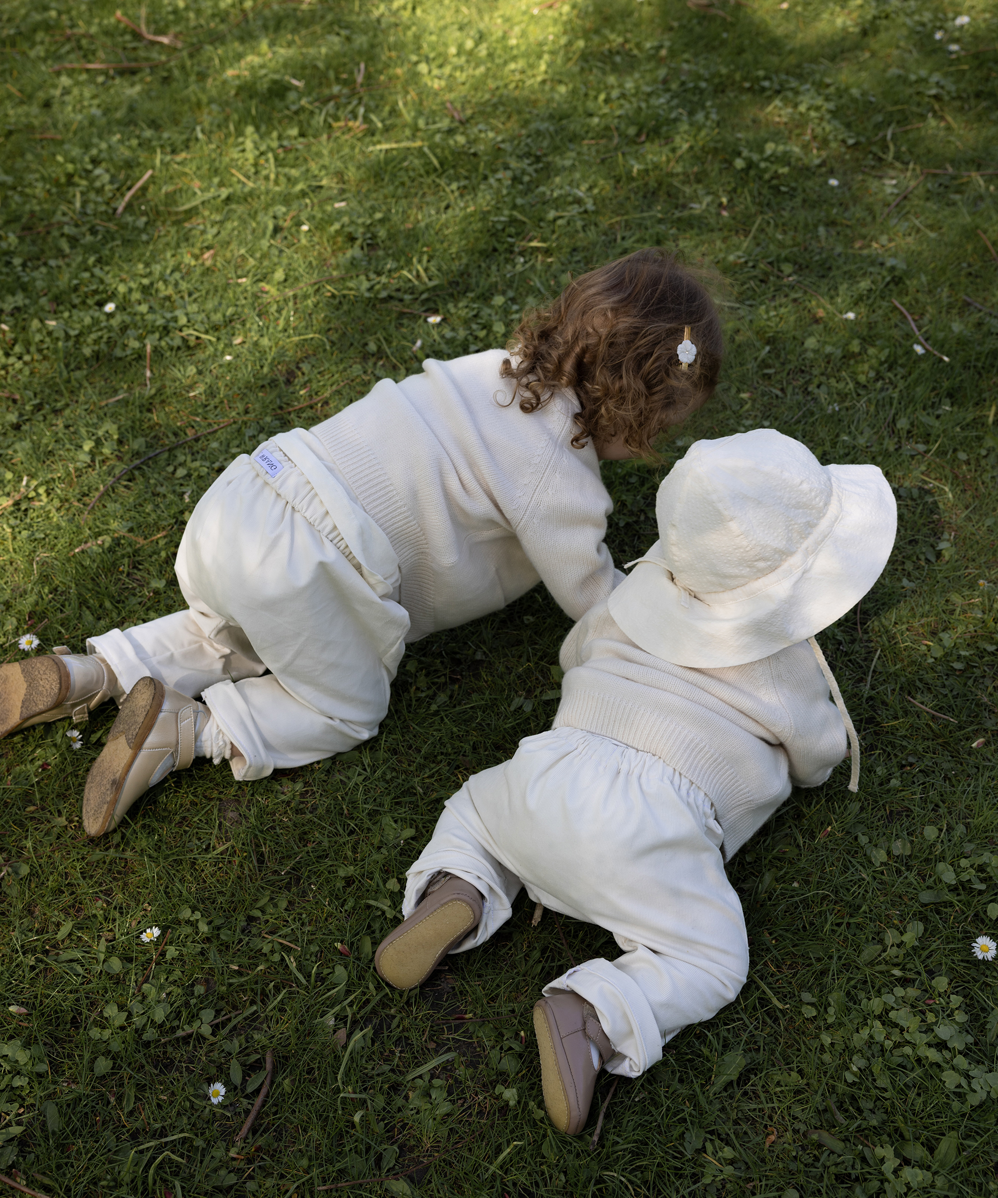 Two young children in matching cream-colored outfits crawl on green grass, enjoying an outdoor setting. Captured from behind, their playful moment is perfect for a Mothers Day memory—one with a clip in her curls, the other sporting a wide-brimmed hat.