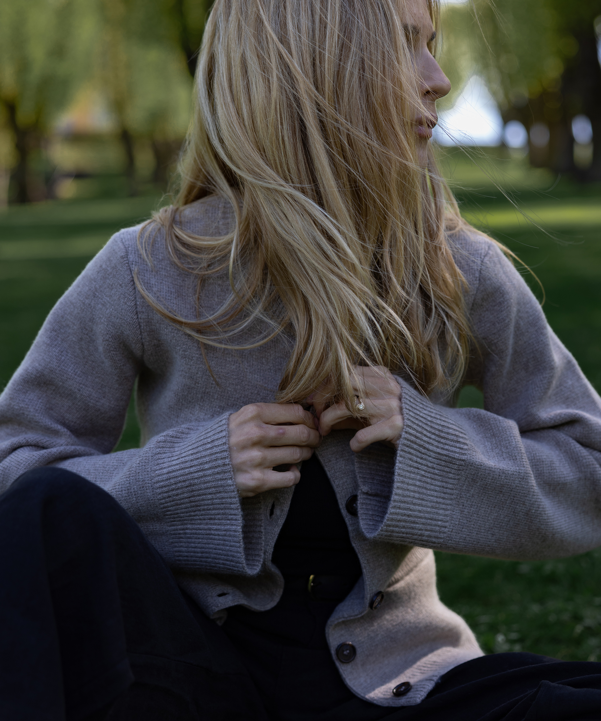 A person with long, blonde hair sits on green grass outdoors, wearing a grey buttoned cardigan and dark pants, gazing to the side with hands near the cardigan buttons—capturing a peaceful Mothers Day moment among trees and sunlight.