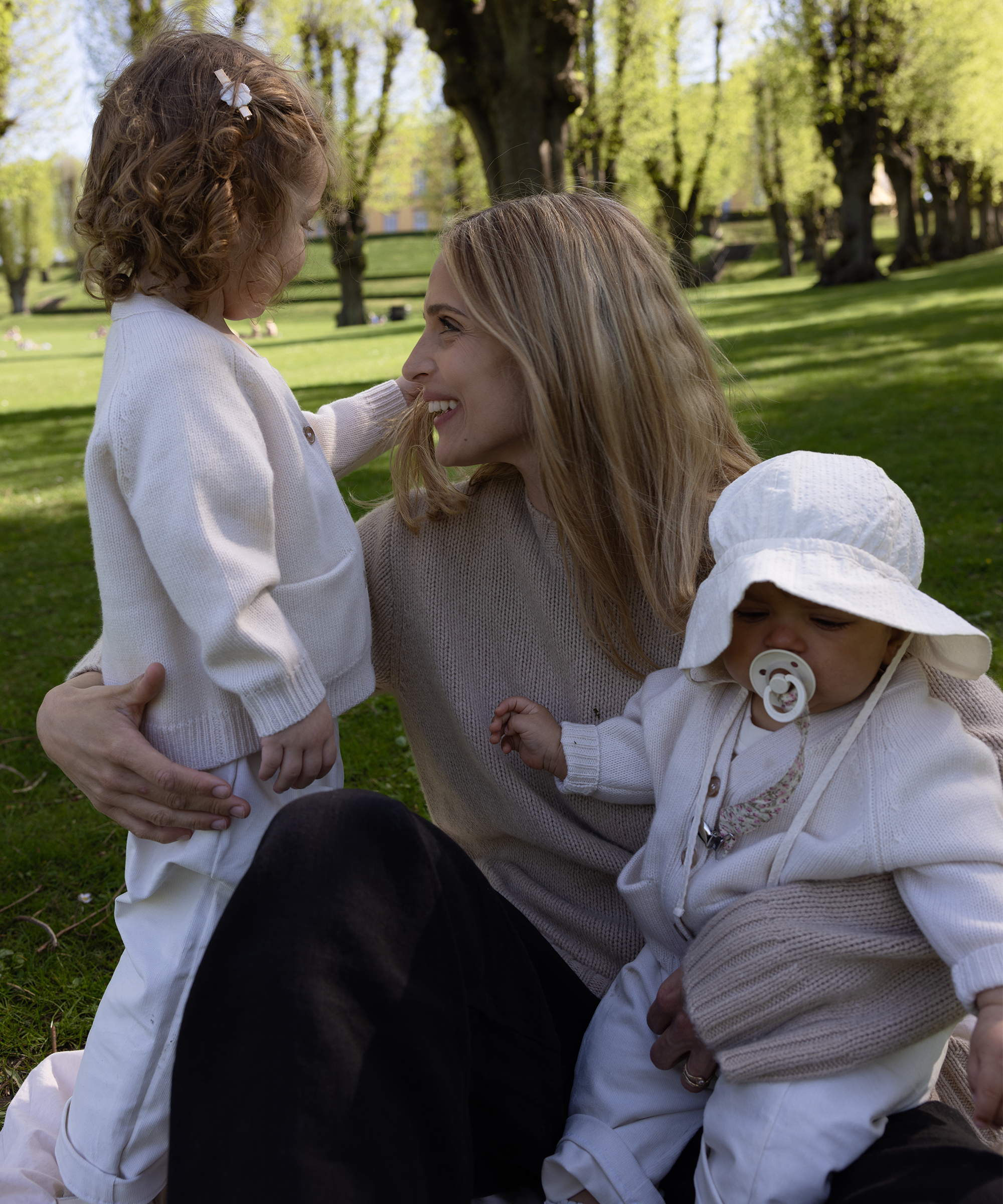 A woman sits on the grass outdoors, smiling at a young child beside her while holding a baby with a pacifier. Surrounded by green trees, this bright scene captures the warmth and joy of Mother’s Day.