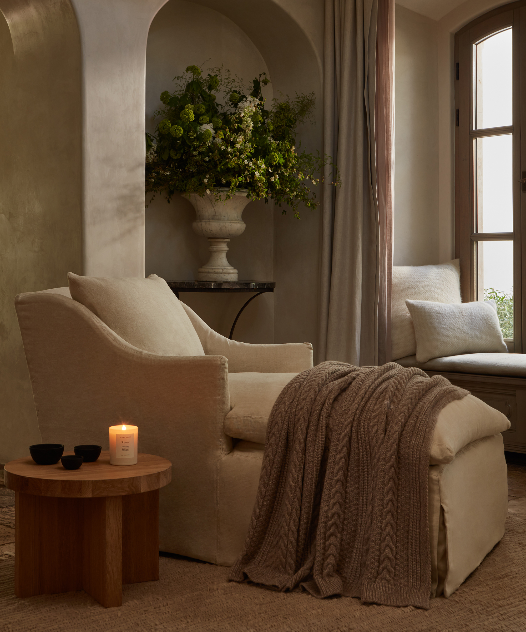 A cozy living space styled by Chloe Crane Leroux features a beige armchair and ottoman draped with a knitted throw, a wooden side table with a lit candle, bowls, greenery in a vase, and a cushioned window seat by an arched window.