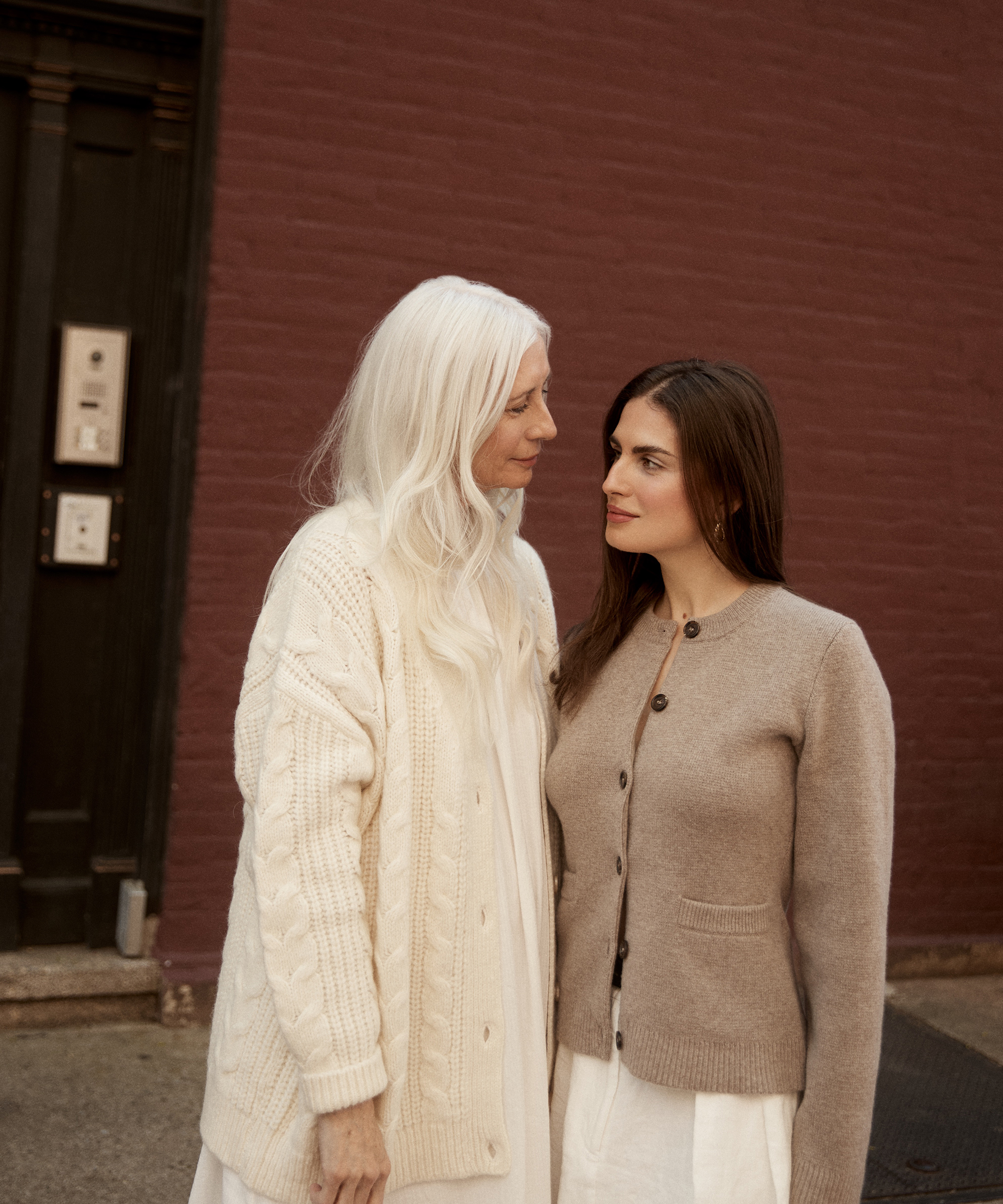 a mother and daughter standing next to each other in front of a brick wall wearing an ivory cardigan and taupe cardigan