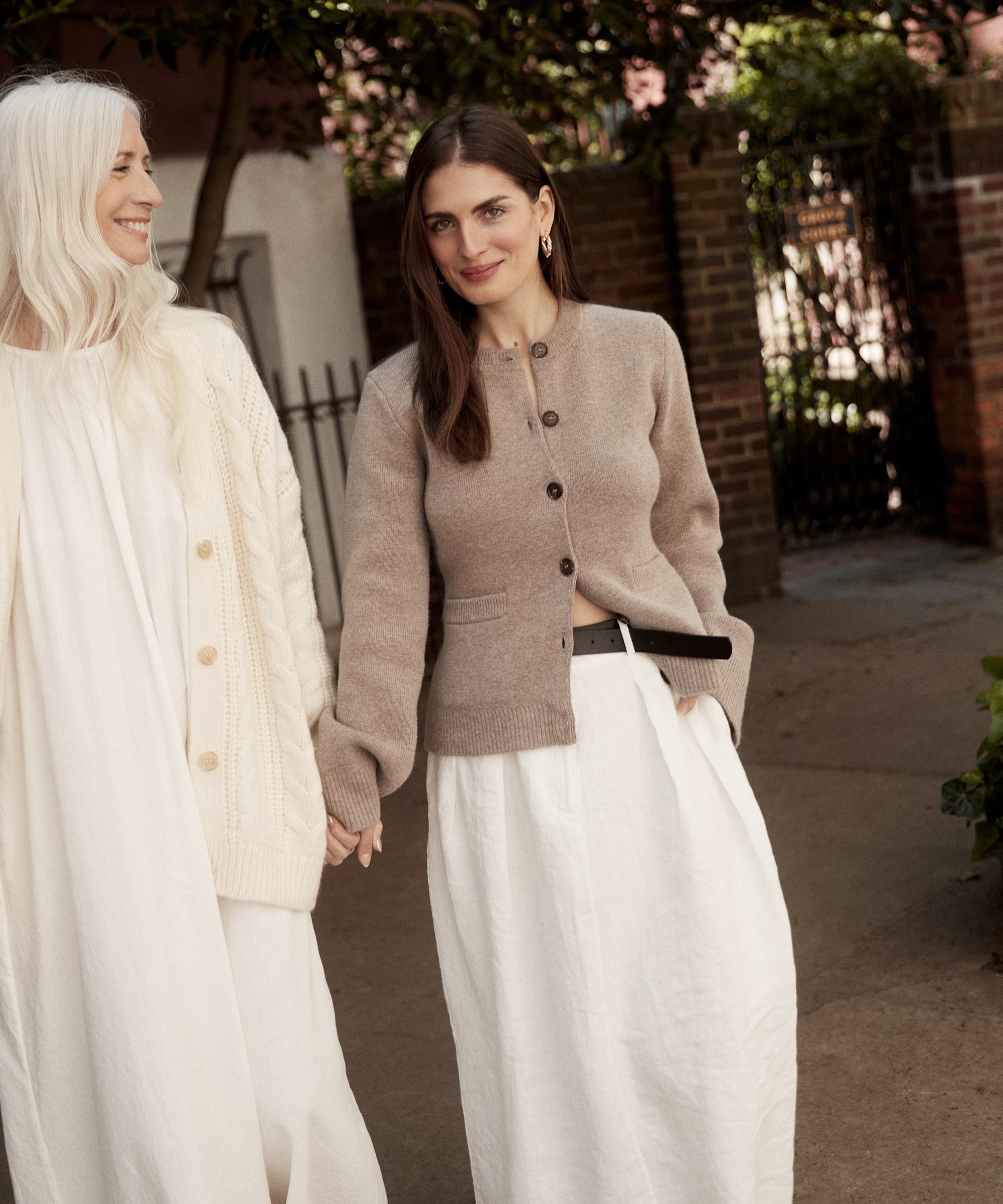 Two women stand outdoors, holding hands and smiling in a scene captured by Chloe Crane Leroux. One wears a cream dress with a chunky knit cardigan; the other, a beige cardigan and white skirt. Greenery and brick walls form the backdrop.