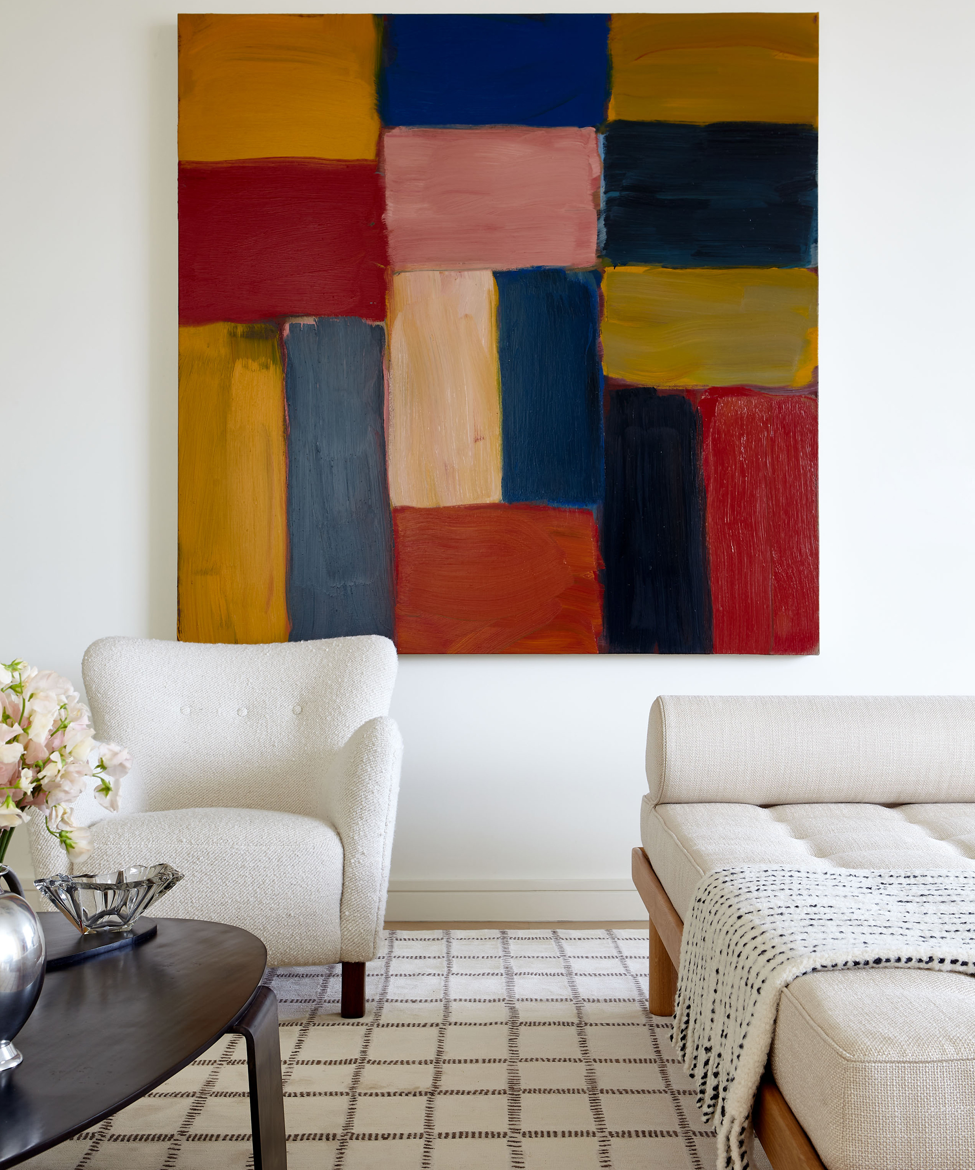 A modern living room with a white armchair, cream sofa, checkered rug, and a large abstract painting featuring colorful rectangular shapes in a cochineal design. A round coffee table and a vase with flowers add finishing touches.