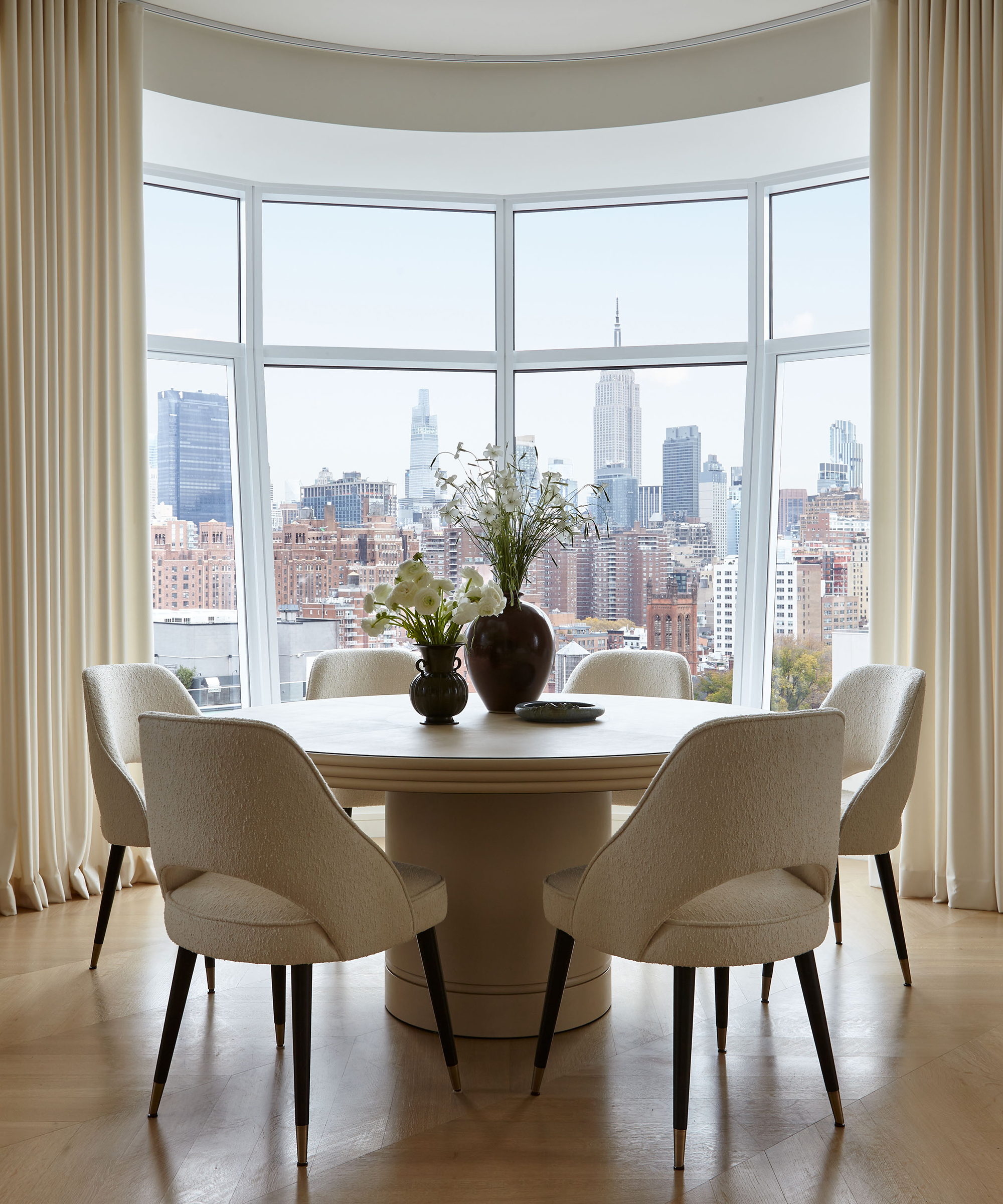 a dining room with a leather round dining table and ivory dining chairs facing floor to ceiling windows of the new york skyline