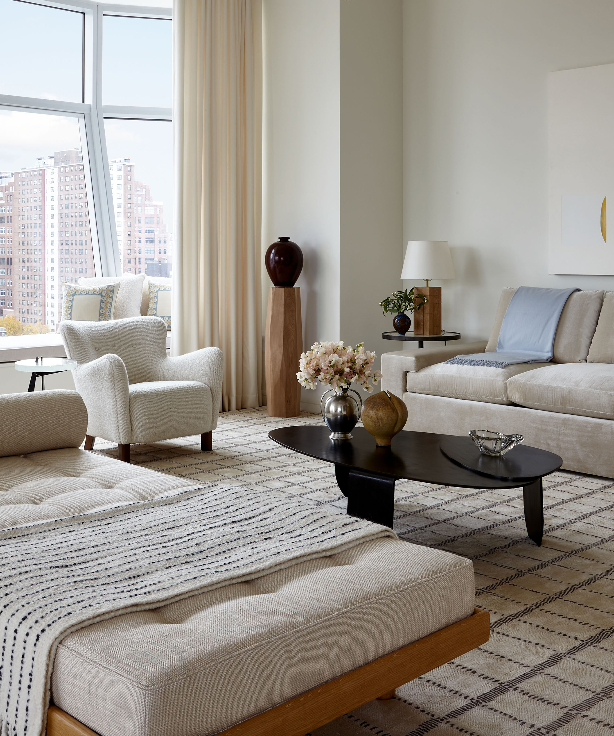 A modern living room with neutral-toned furniture and cochineal design accents, featuring a sofa, armchair, daybed, black coffee table with decor, large windows offering city views, and light curtains.
