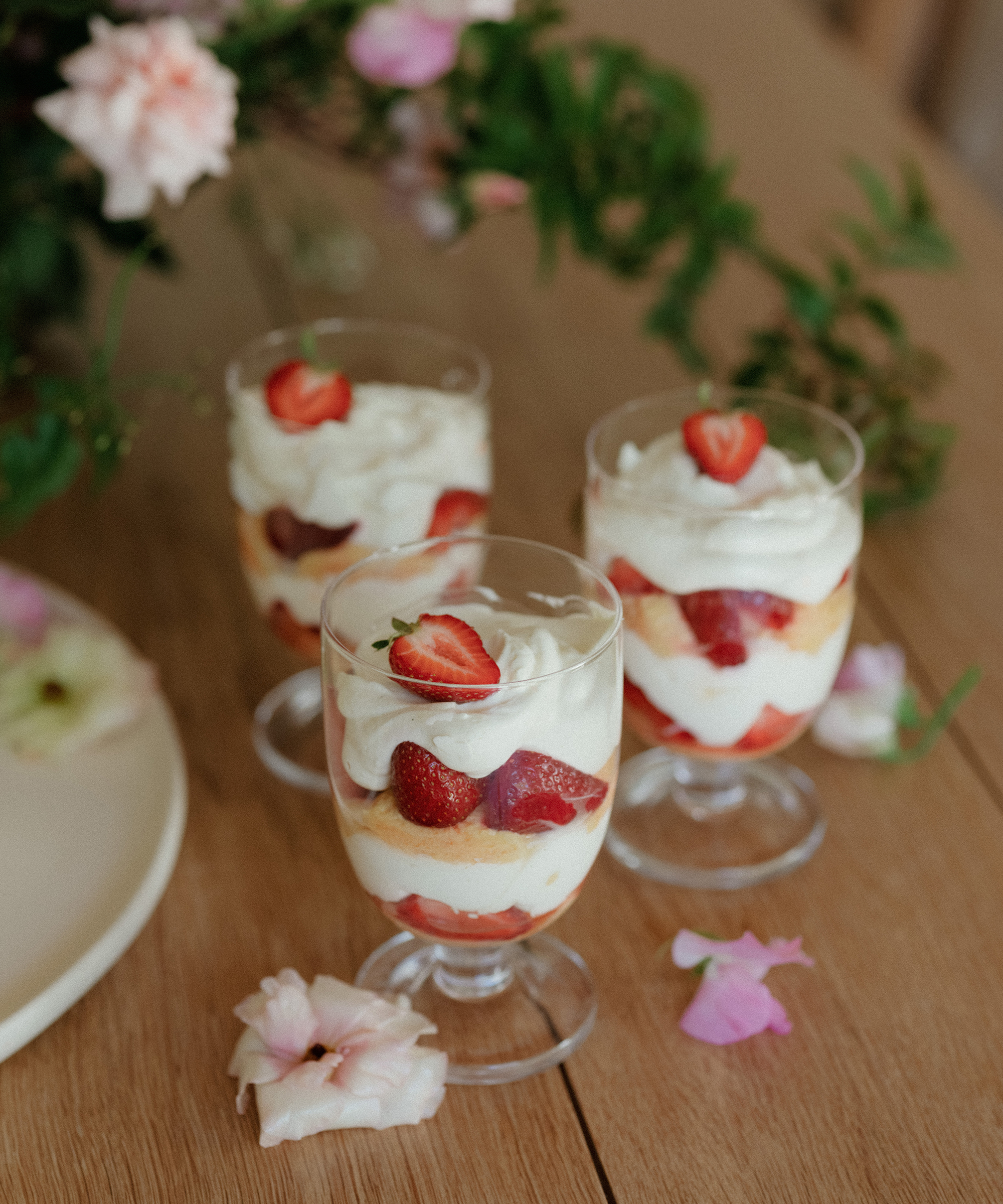 Three glass dessert cups filled with layers of whipped cream, sponge cake, and strawberries are arranged on a wooden table, surrounded by pink flowers and greenery—perfect inspiration for your next dessert recipes.