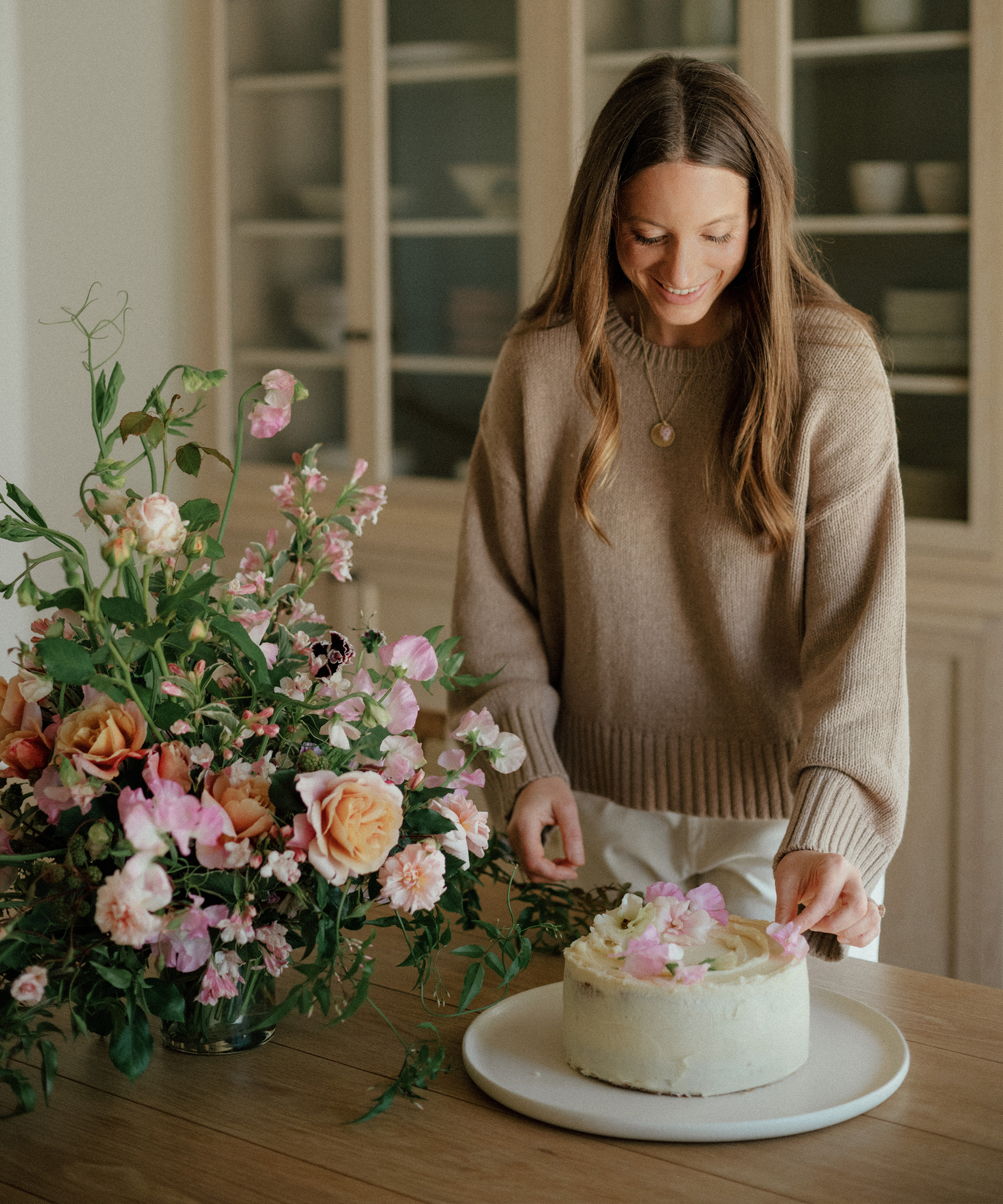 danielle in a taupe sweater decorating a iced carrot cake on a white serving platter on a wood dining table with flowers