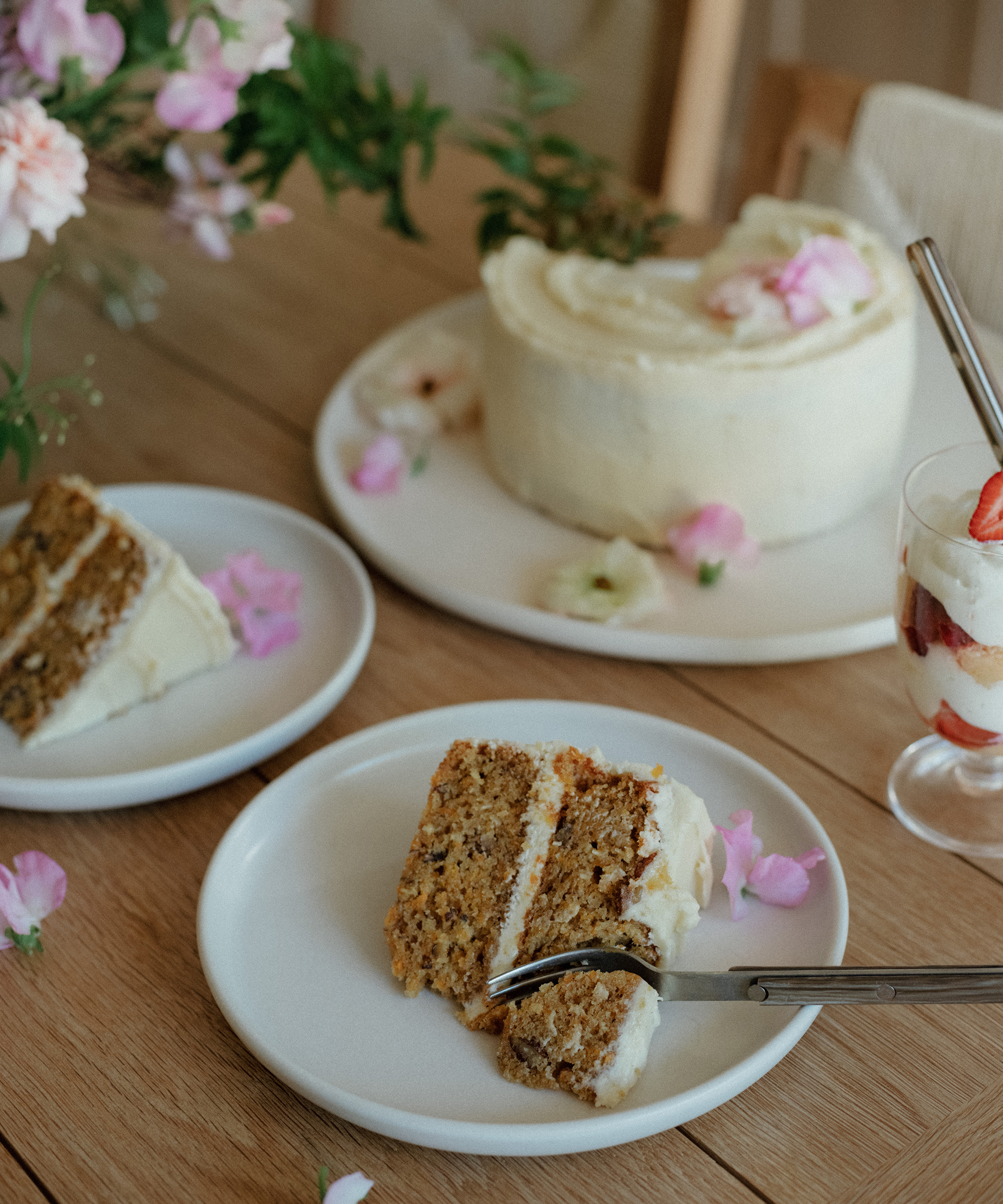 A slice of layered carrot cake with cream cheese frosting sits on a white plate with a fork, beside a whole cake and a dessert glass with strawberries—an inviting scene for anyone who loves dessert recipes, all adorned with pink flower petals on a wooden table.