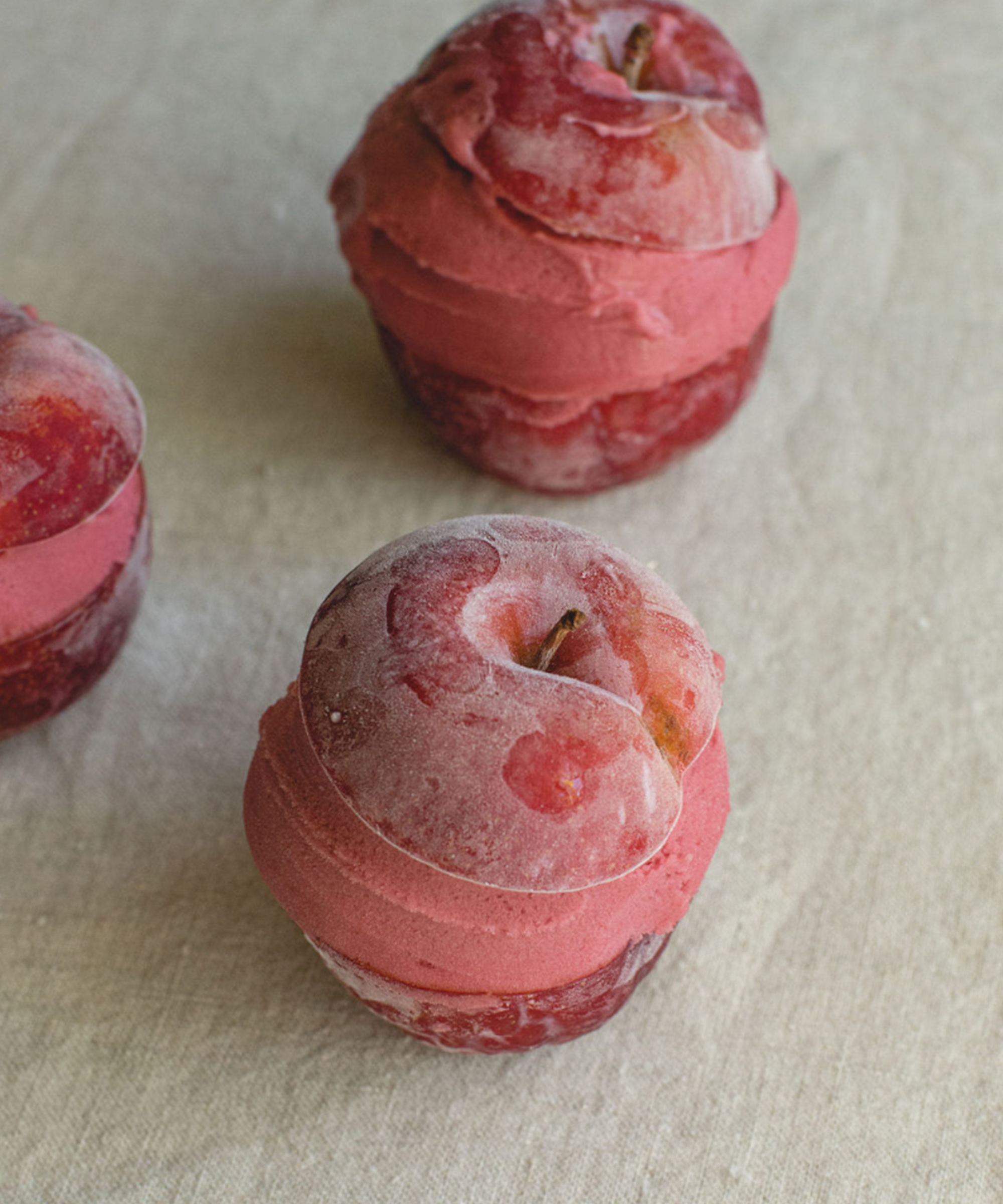 Three whole plums are sliced horizontally and filled with a layer of pink sorbet, creating a delightful dessert that resembles ice cream sandwiches, displayed on a light-colored fabric surface.