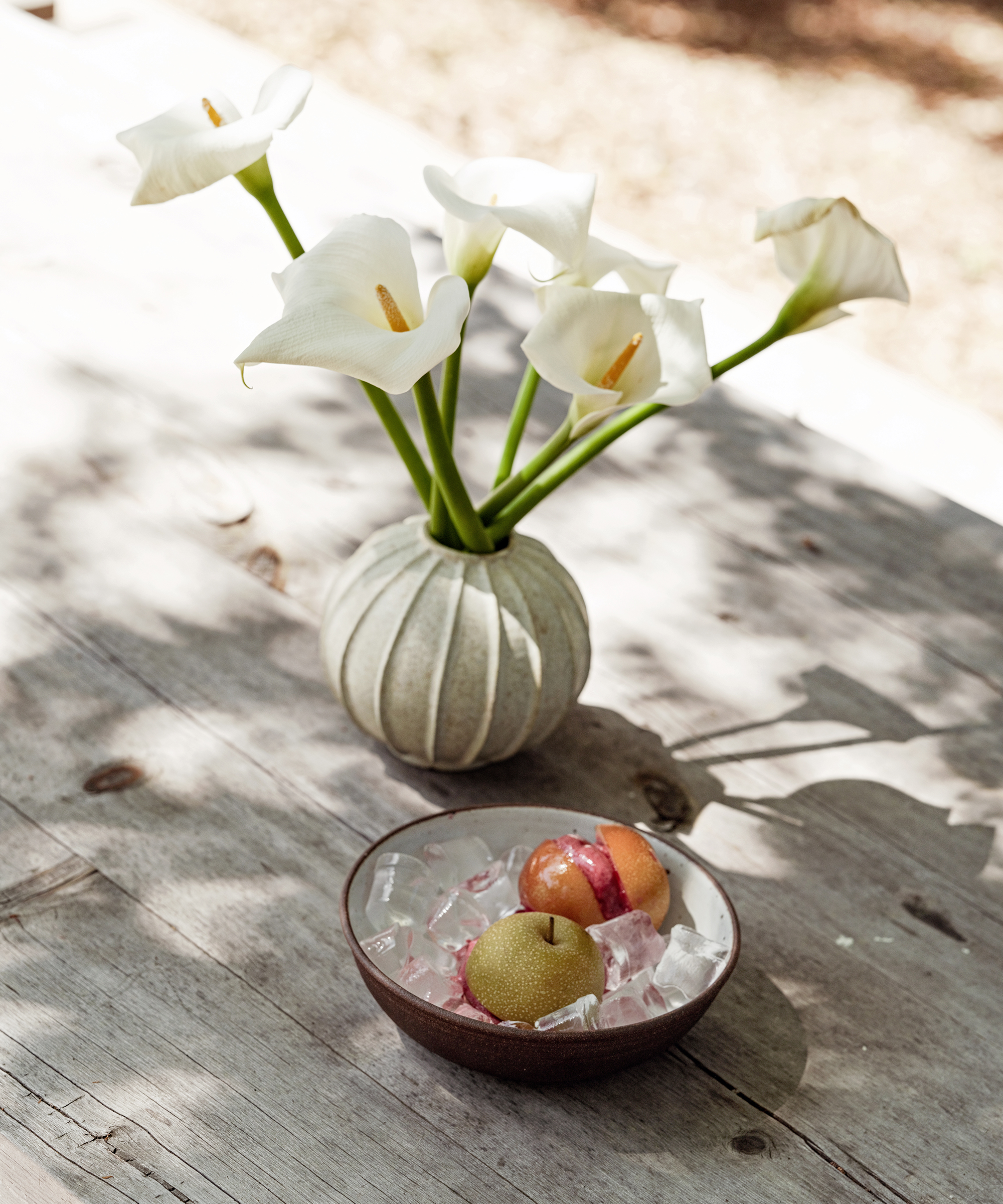 a wood outdoor dining table with a round bowl filled with ice and pears stuffed with sorbet next to a creme vase with flowers
