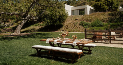 an outdoor dining table and dining bench in the grass in front of a house and horse stable