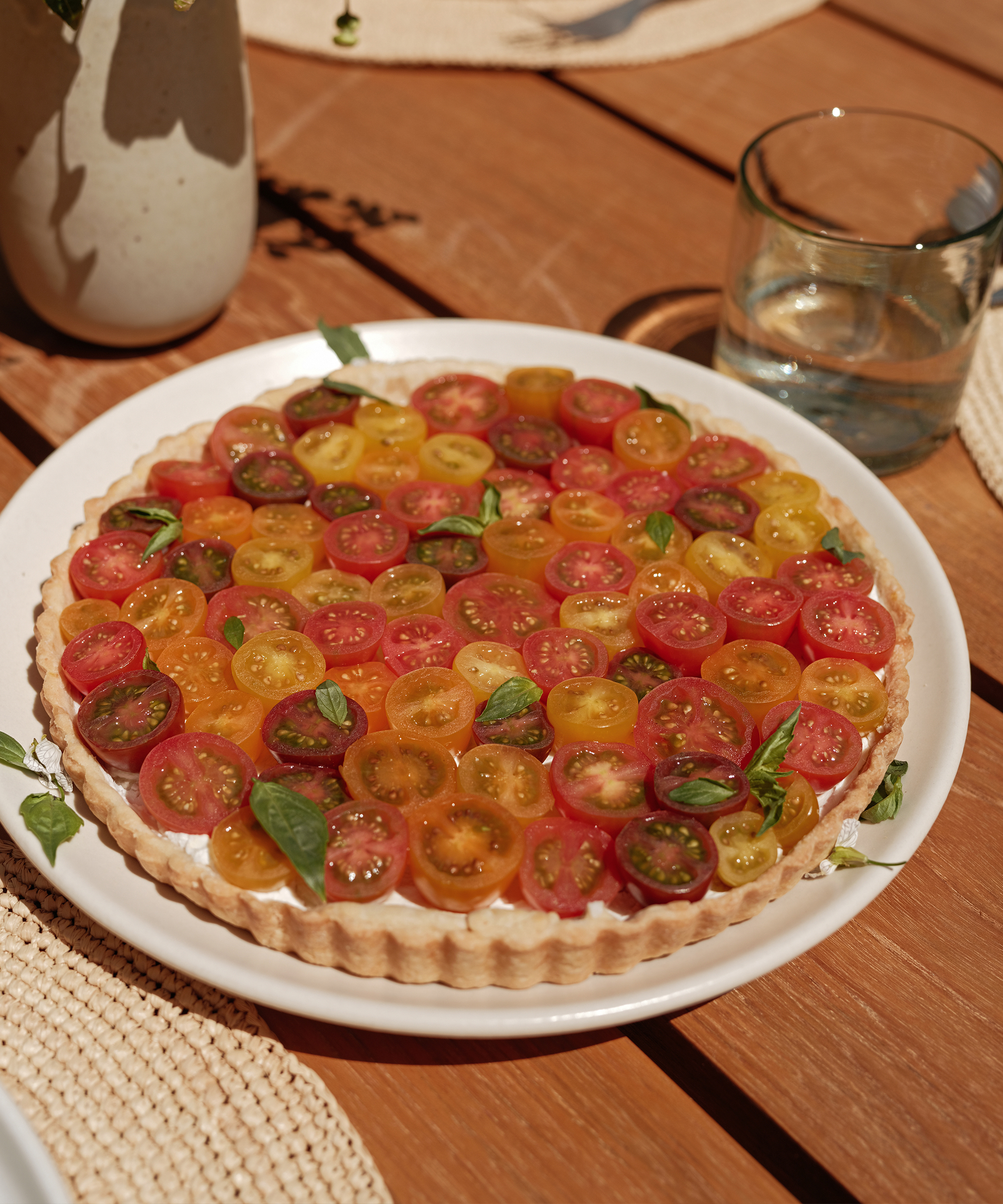 A tart topped with sliced red, yellow, and green cherry tomatoes, garnished with basil leaves, sits on a white plate on a wooden table next to a glass of water—perfect for your collection of brunch recipes.