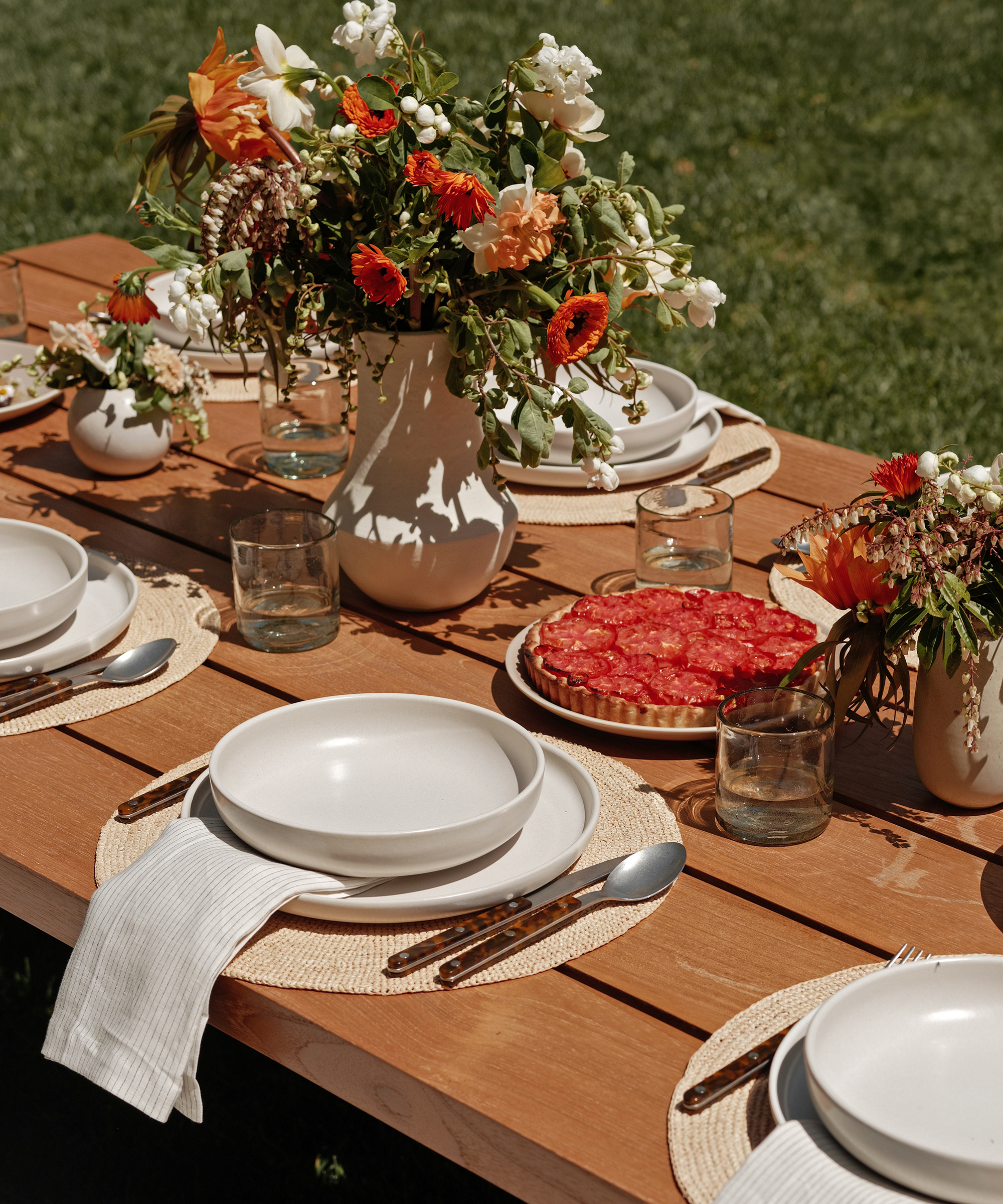 A wooden outdoor table set for brunch recipes, featuring white plates, woven placemats, glasses, and cutlery. White cloth napkins and vases of vibrant flowers add charm, while a platter of sliced tomatoes sits atop the table with grass in the background.