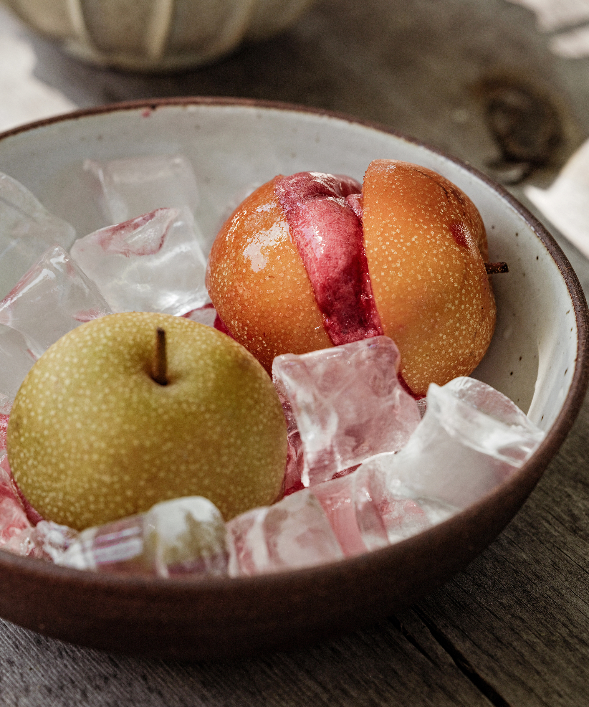 A bowl with ice cubes holds an Asian pear and a sliced plum, creating a refreshing dessert display on a rustic wooden surface.