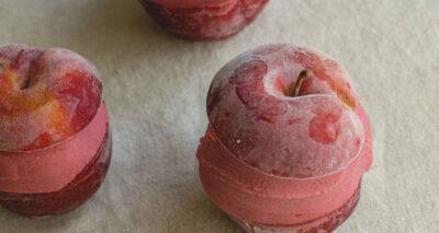 Three plums with a layer of pink ice cream sandwiched in the middle rest on a light-colored surface. Slightly frosted, they create a unique chilled dessert with an inviting, sweet appearance.