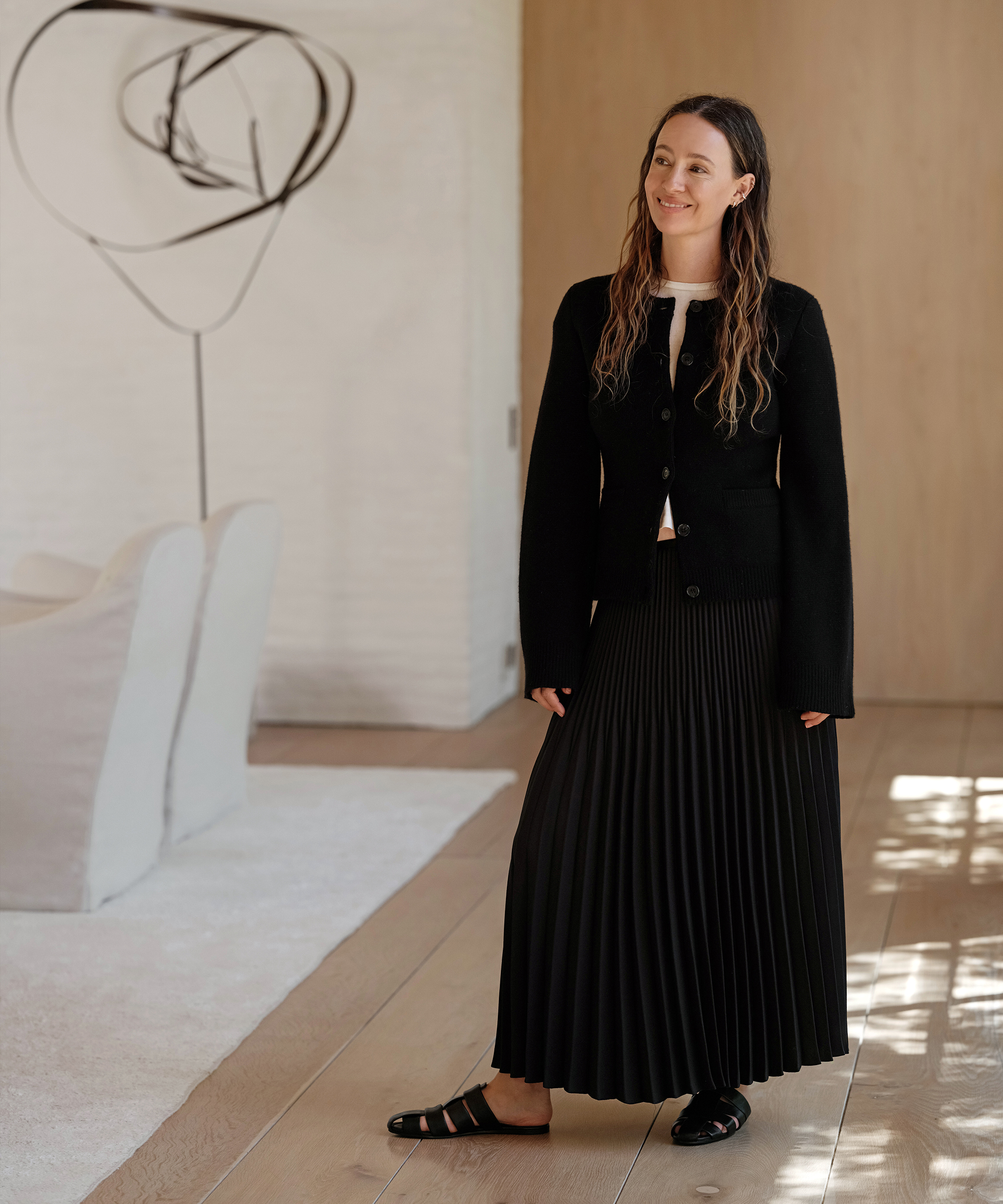 jenni wearing a black cardigan and black pleated skirt with black mules standing inside her living room