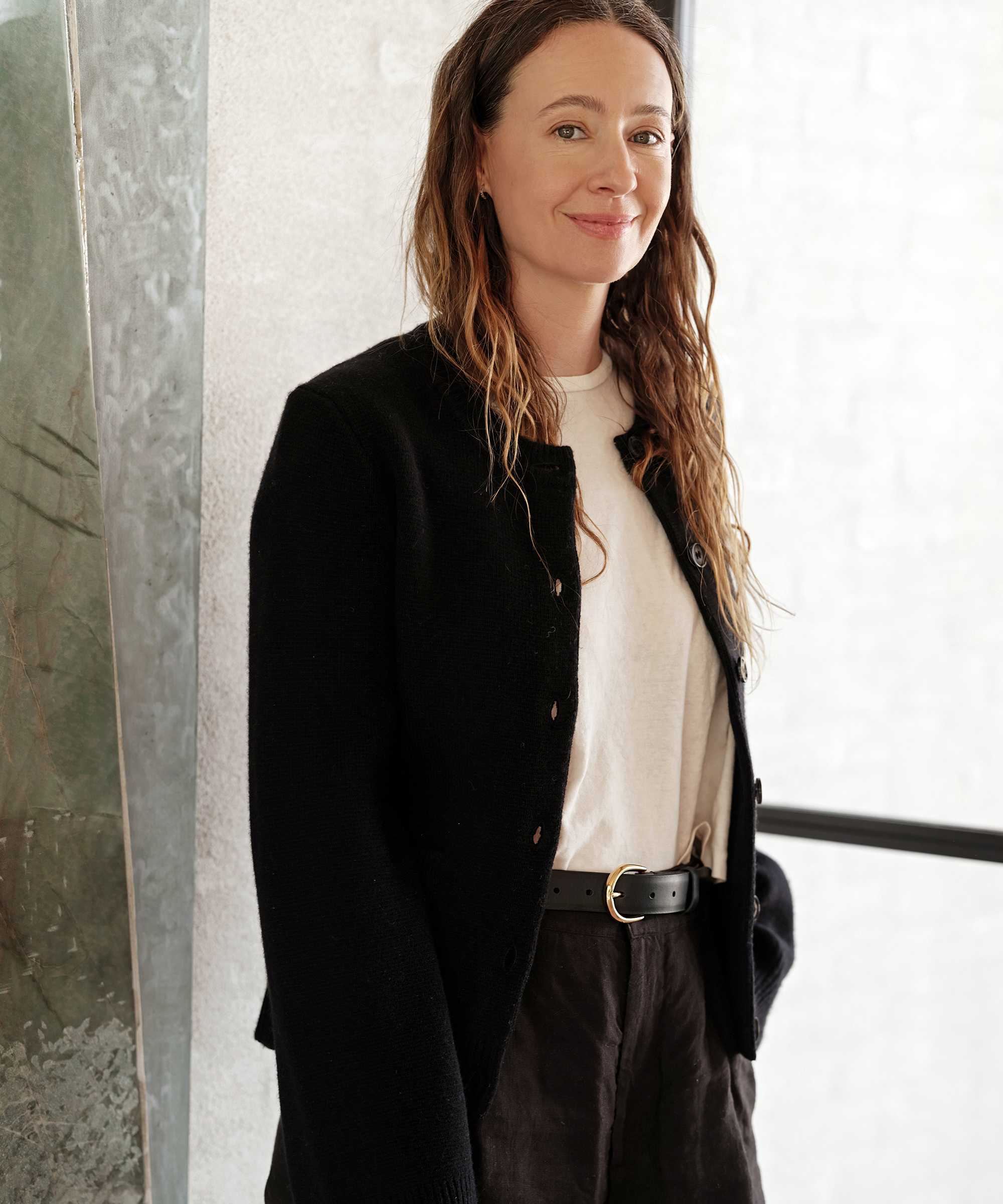A woman with long, wavy brown hair stands indoors, smiling gently. She is wearing a black jacket over a white t-shirt, black belt, and dark pants, with one hand in her pocket. She stands near a large window.