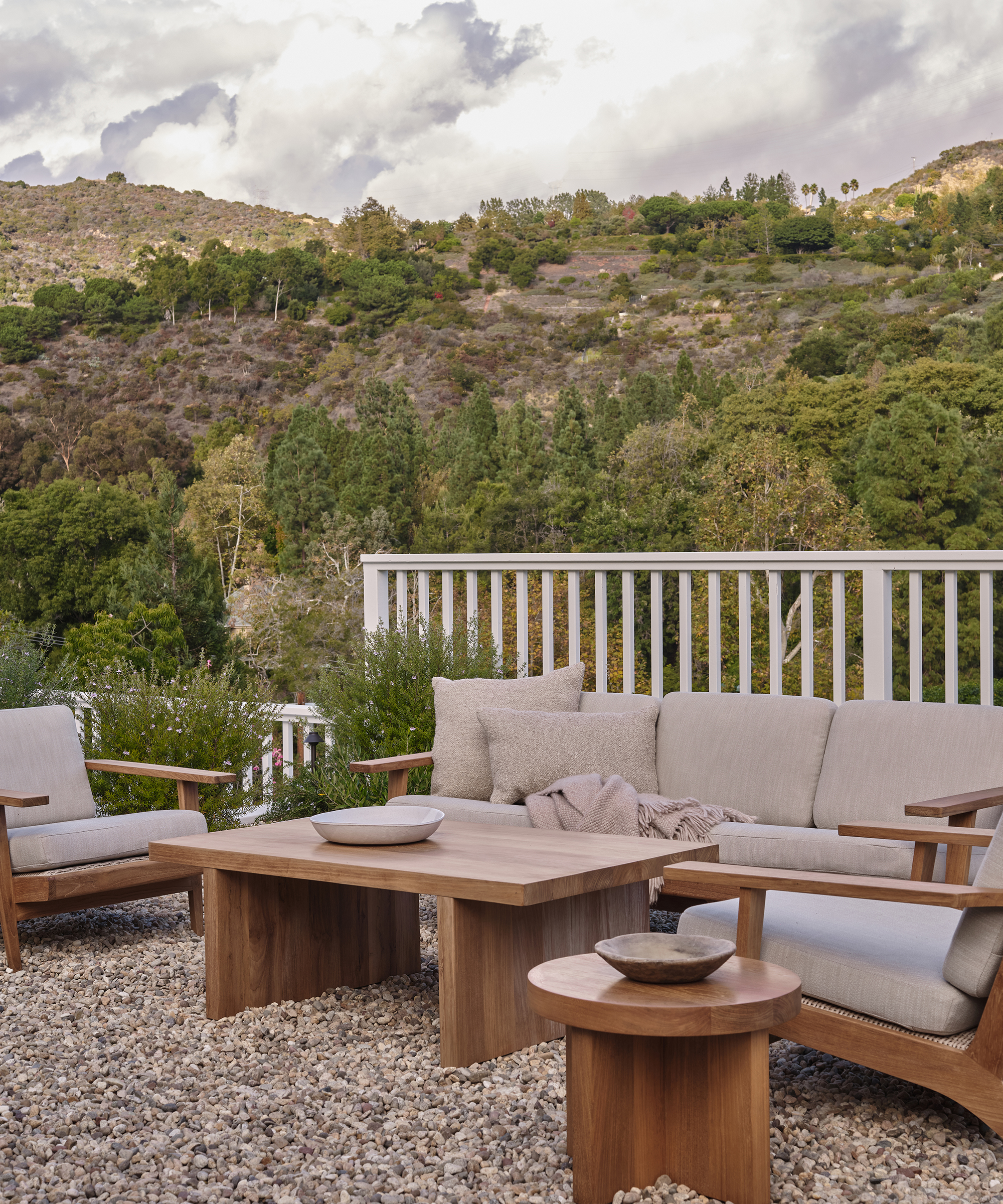 A modern outdoor patio with stylish cushioned wooden outdoor furniture and tables sits on gravel, surrounded by lush greenery and mountains under a cloudy sky.