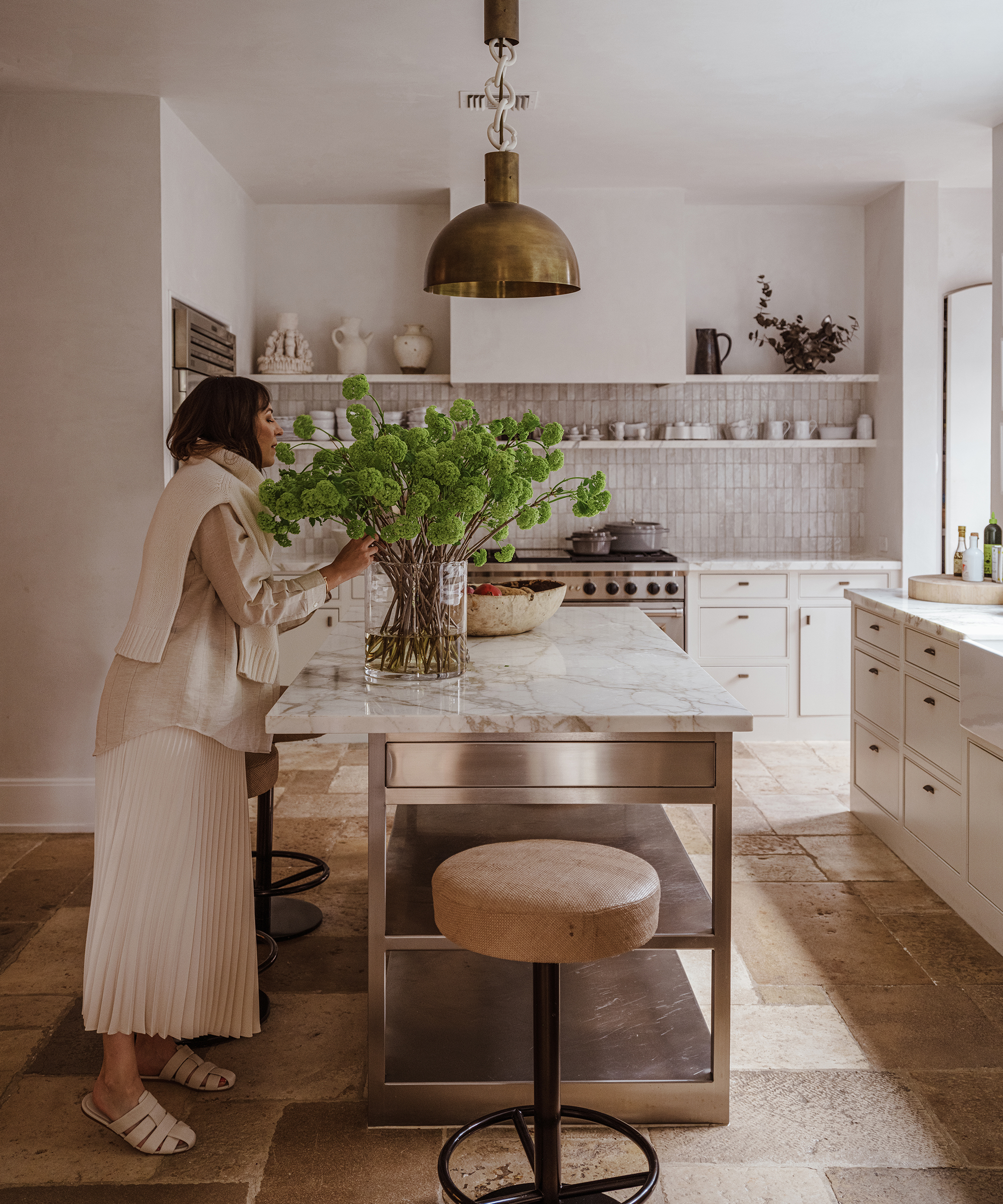 Margaret Naeve arranges green flowers in a large vase on a marble kitchen island in a bright, modern kitchen with white cabinets, open shelves, and warm natural lighting.