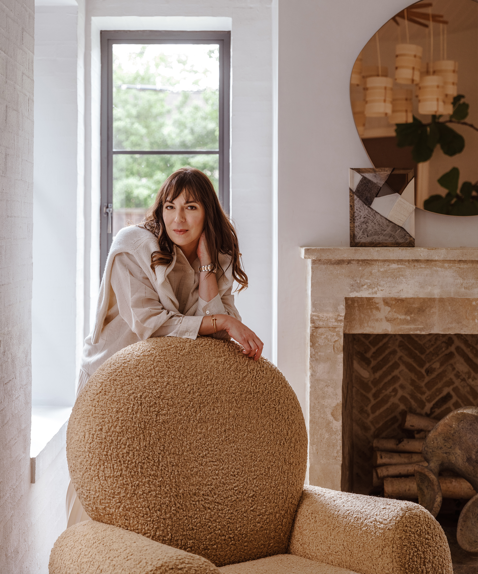 A woman with long brown hair leans on a round, textured beige chair in a cozy, light-filled room designed by Margaret Naeve, featuring a stone fireplace, a round mirror, and a window framing lush greenery outside.
