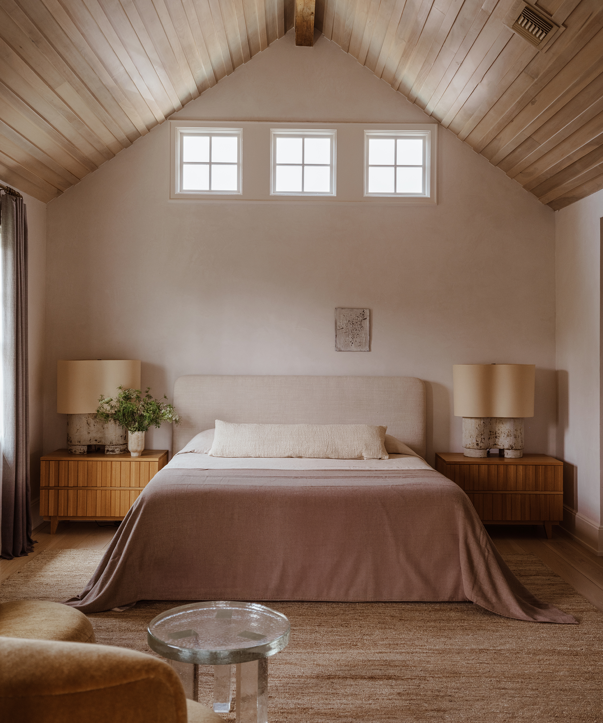 A cozy bedroom with a vaulted wood ceiling, neutral-toned walls, and a bed with a taupe blanket. Two Margaret Naeve nightstands with lamps flank the bed, while soft natural light fills the room.