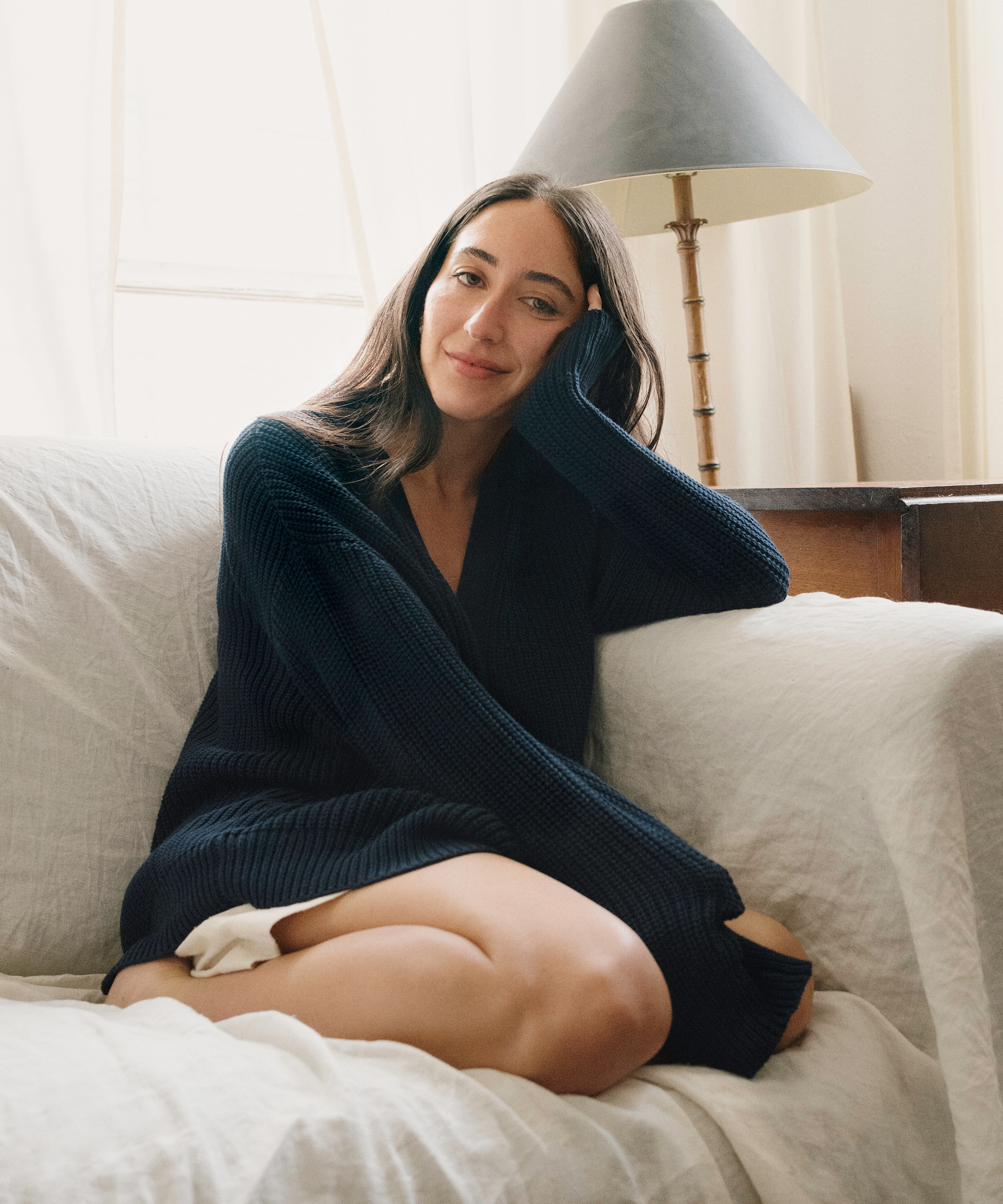 Alexis Badiyi, with long dark hair, sits on a white couch in an oversized sweater. She rests her head on her hand and smiles softly, with a lamp and sheer curtains in the background.
