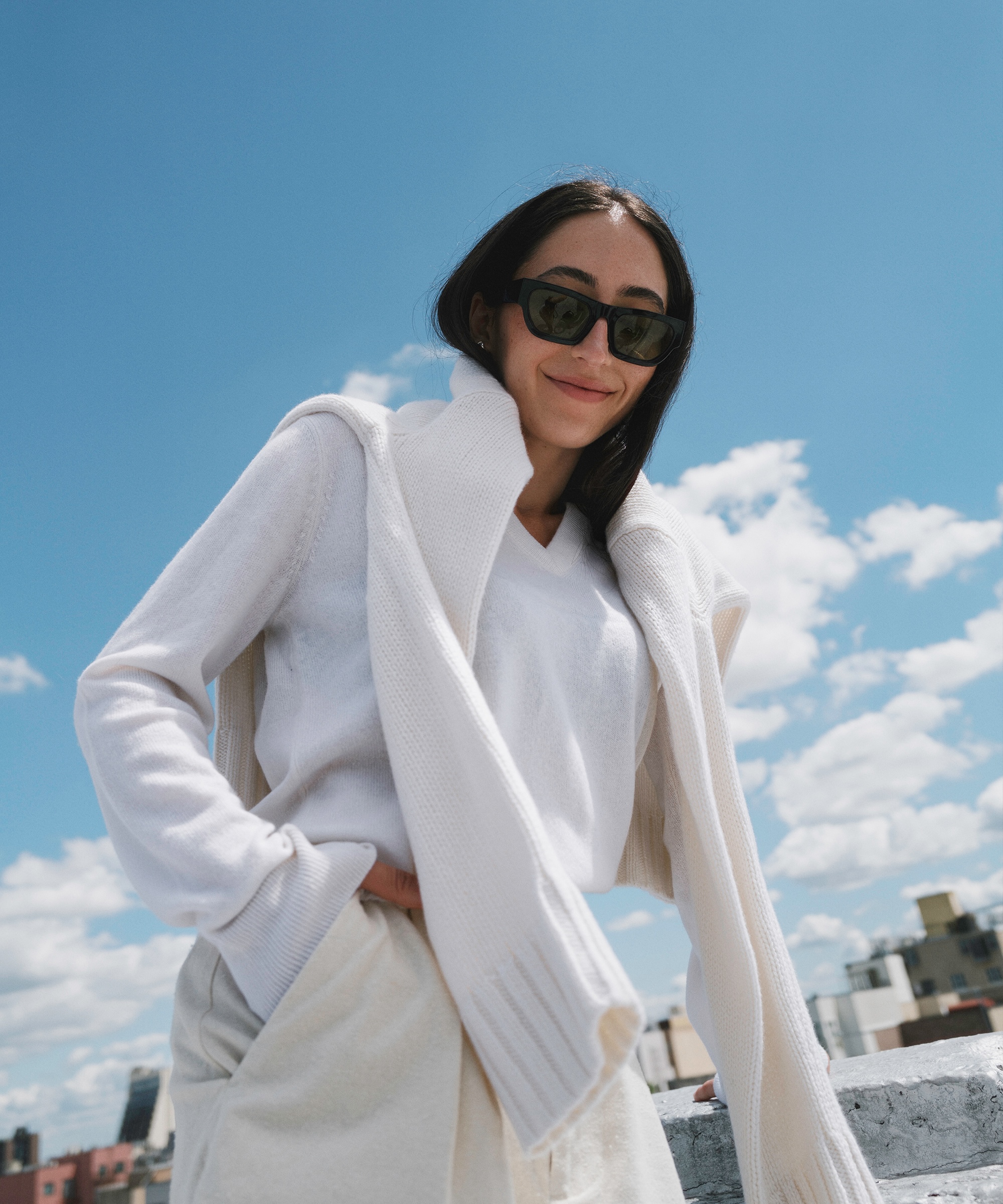Alexis Badiyi, wearing sunglasses and dressed in white, stands outdoors under a blue sky with clouds. She smiles with a sweater draped over her shoulders, while buildings rise in the background.