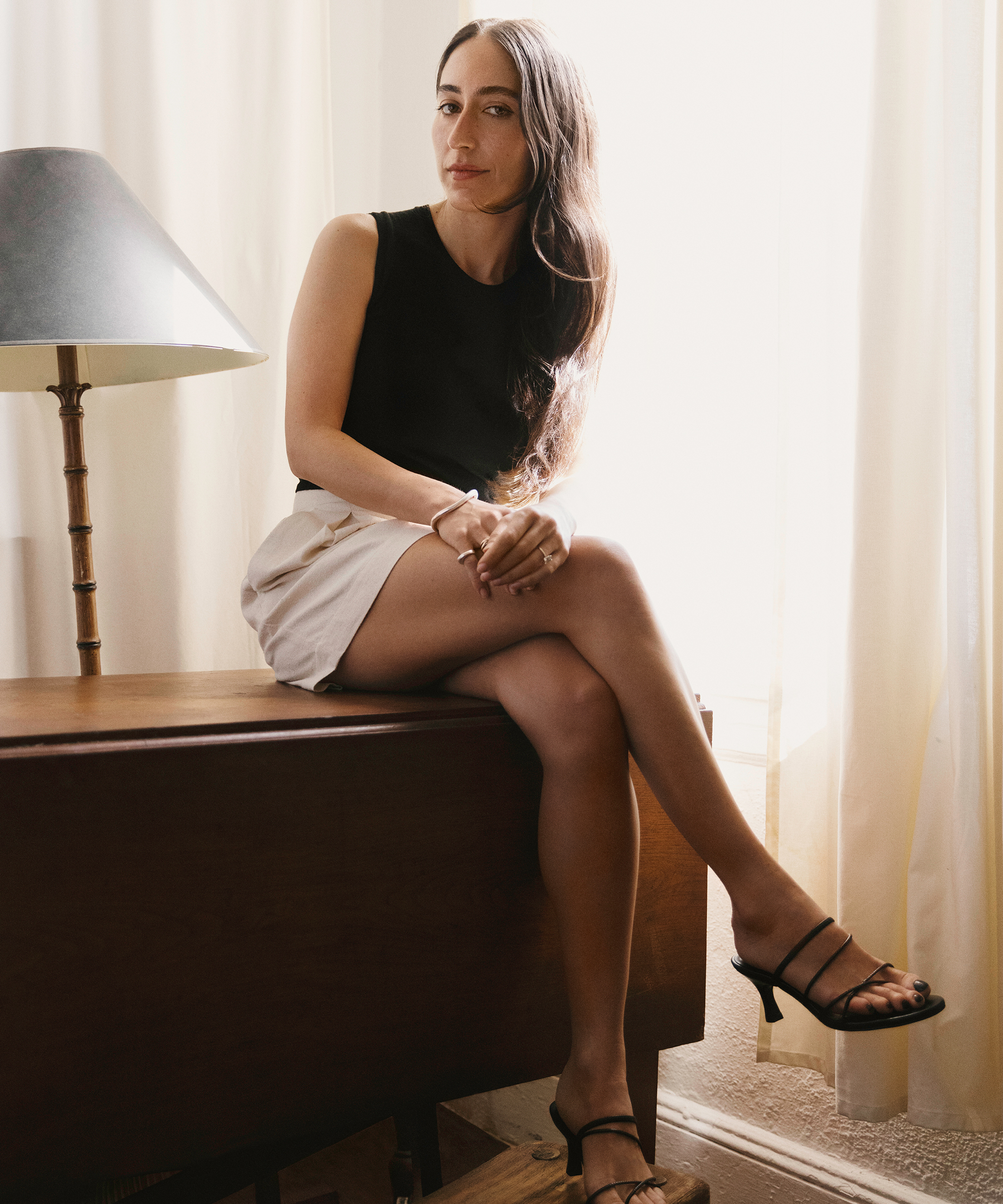 alexis wearing ivory pleated shorts and a black tan sitting on a wooden table next to a window with ivory curtains