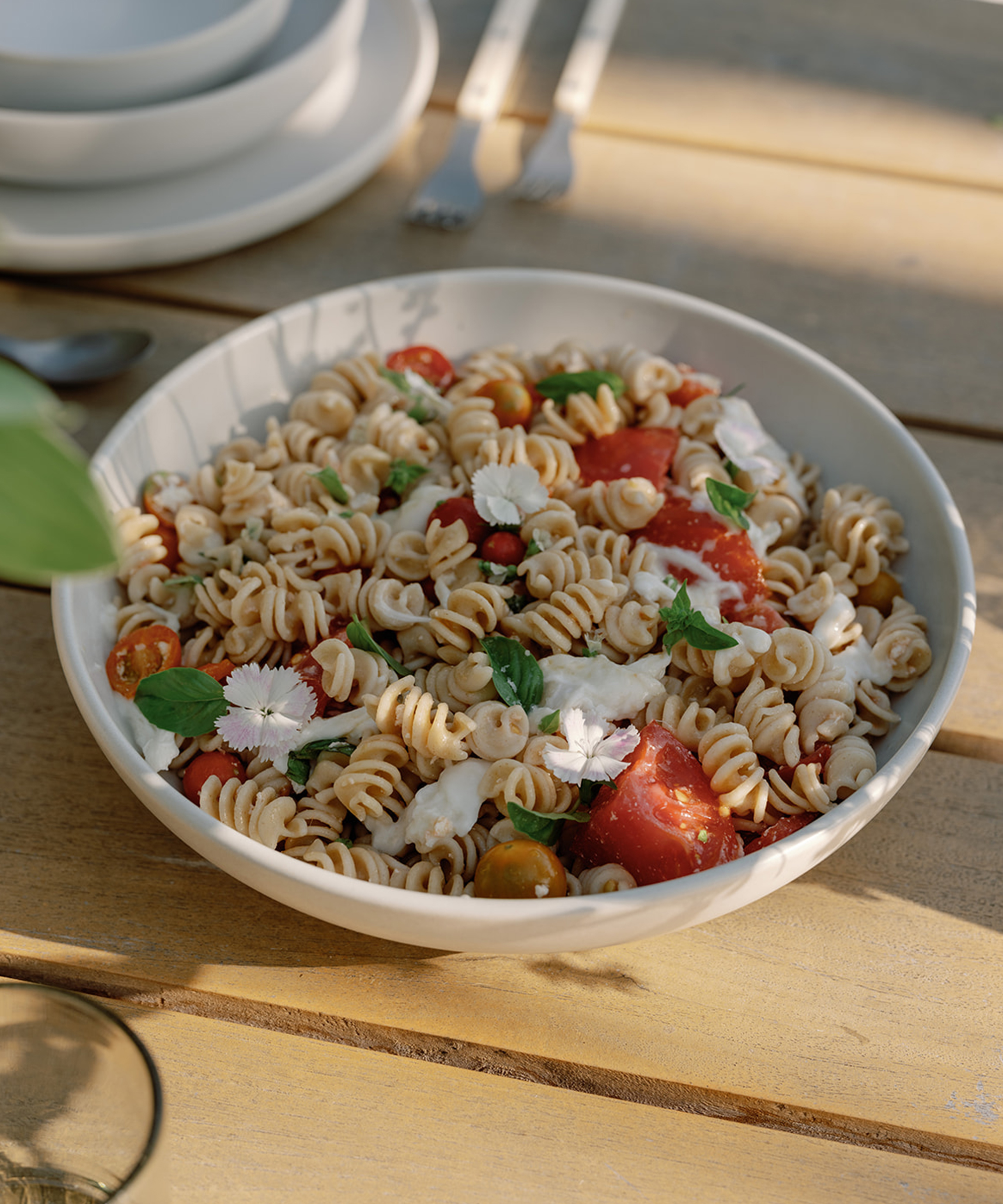 A bowl of rotini pasta salad with cherry tomatoes, mozzarella, fresh basil, and edible flowers sits on a wooden table in sunlight—perfect for a summer dinner—with plates and forks in the background.