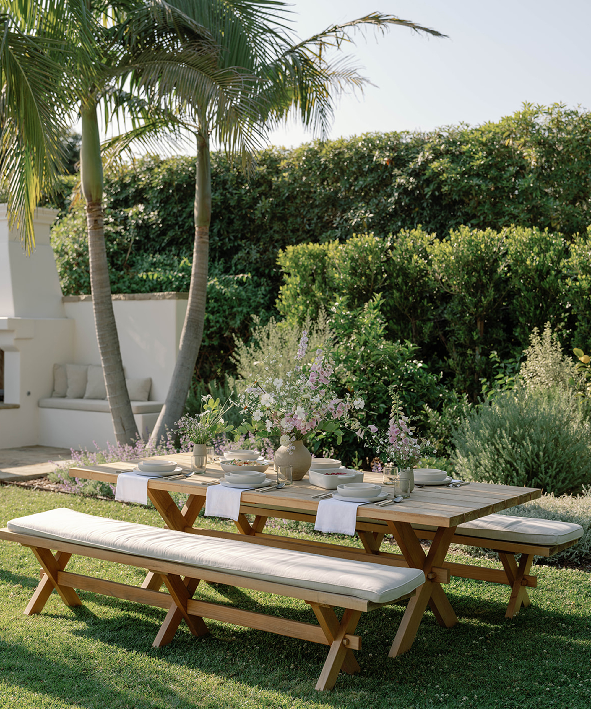 a wooden outdoor dining table with a bench set on green grass under a palm tree
