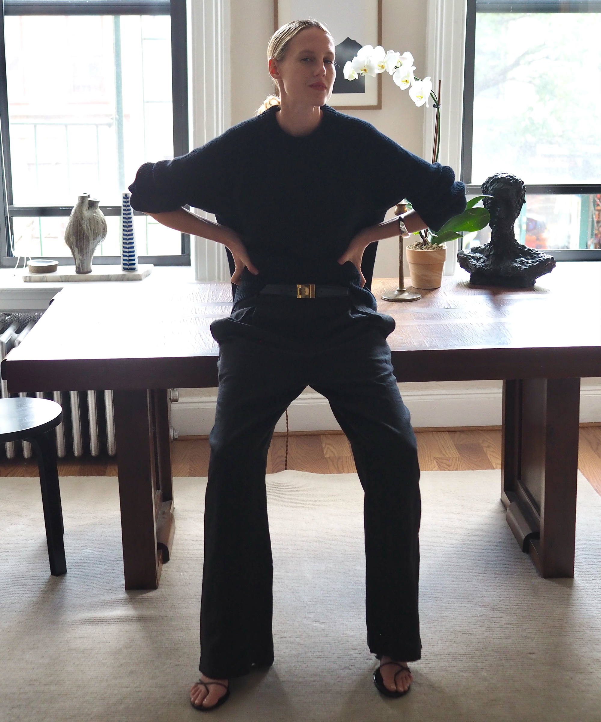 a women leaning on her wooden desk wearing black trousers and a navy cotton fisherman sweater