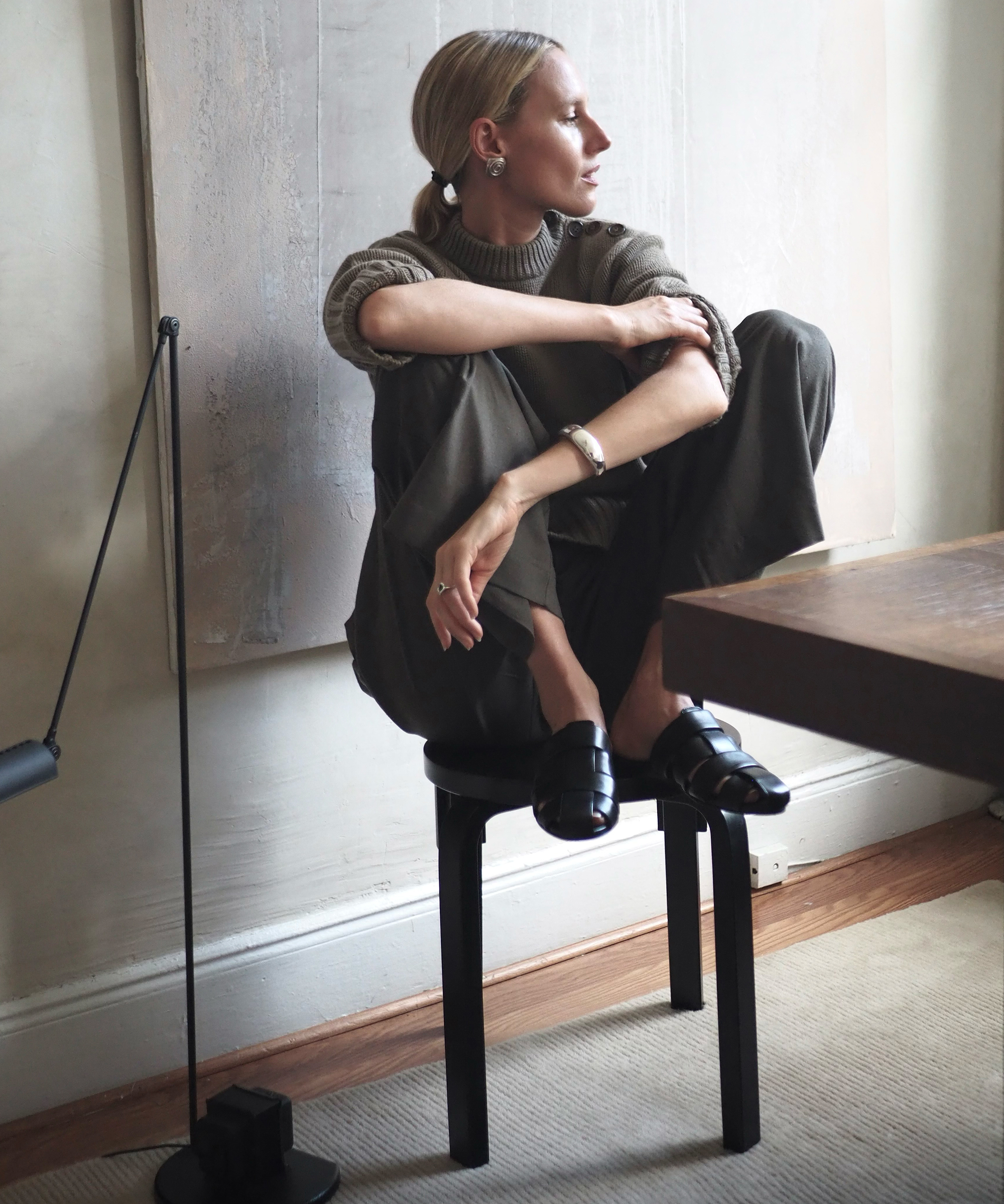 Elly McGaw, with blonde hair, sits cross-legged on a black stool, wearing a brown sweater, dark pants, and black loafers. She looks to the side, arms resting on her knees in a softly lit, modern room.