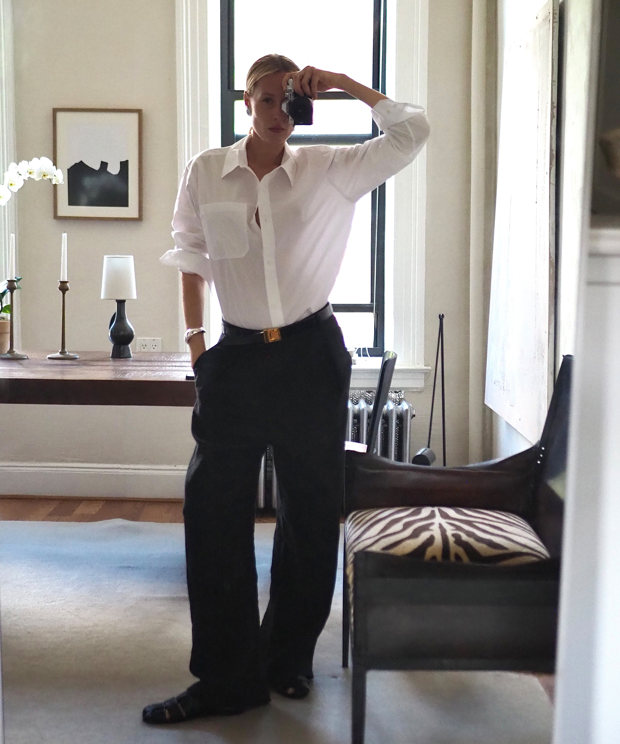 elly mcgaw in black trousers and white button down taking a self portrait in a mirror leaning against her desk