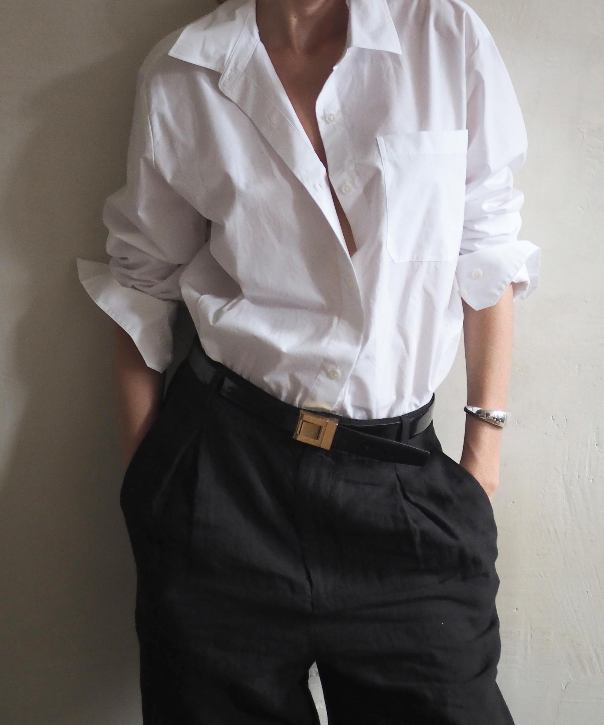 Person wearing an oversized white button-down shirt with rolled-up sleeves, tucked into high-waisted black pants with a belt, standing against a plain light-colored wall. The effortless style is reminiscent of Elly McGaw’s signature look. Hands are in the pockets.