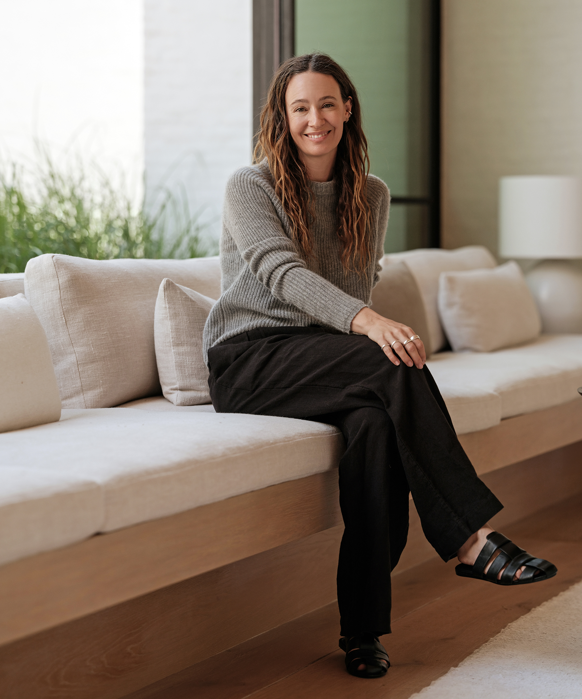 jenni sitting on a built in couch wearing black linen trousers and a grey cashmere fisherman sweater with black mules