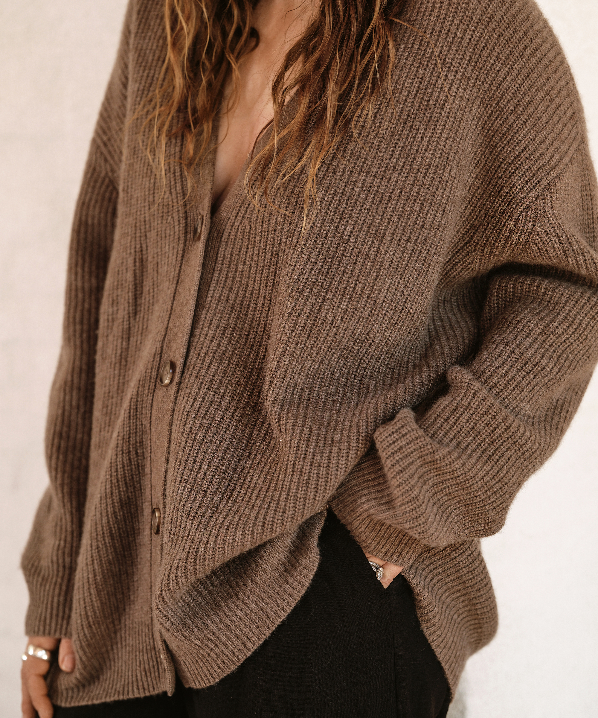 A person wears a loose, brown, ribbed cocoon cardigan with large buttons and black pants. Long wavy hair is partially visible, hand tucked in their pocket, with their face not shown.