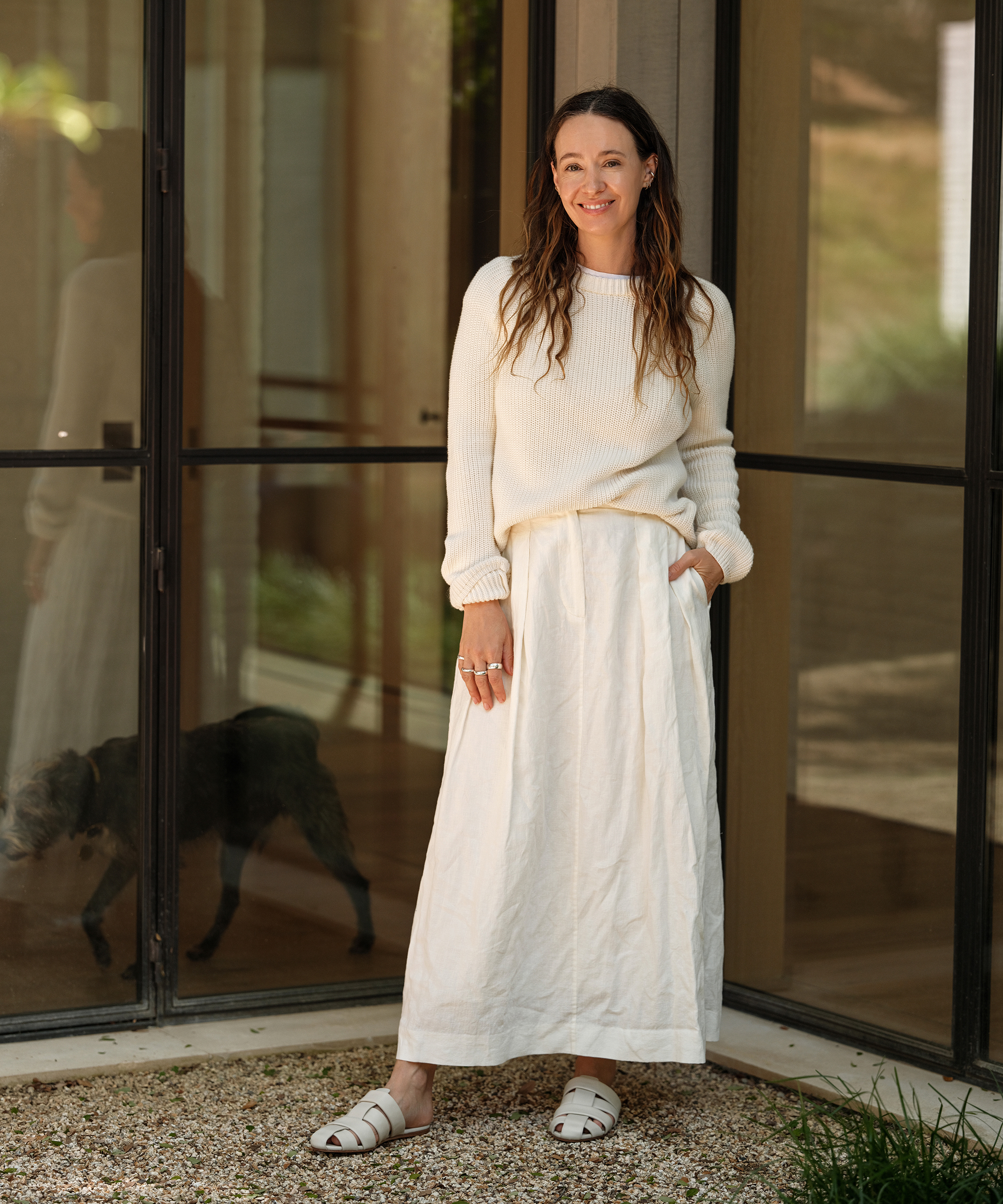 jenni standing outside in front of a glass door wearing a white linen maxi skirt and an ivory cotton fisherman sweater with mules that are ivory