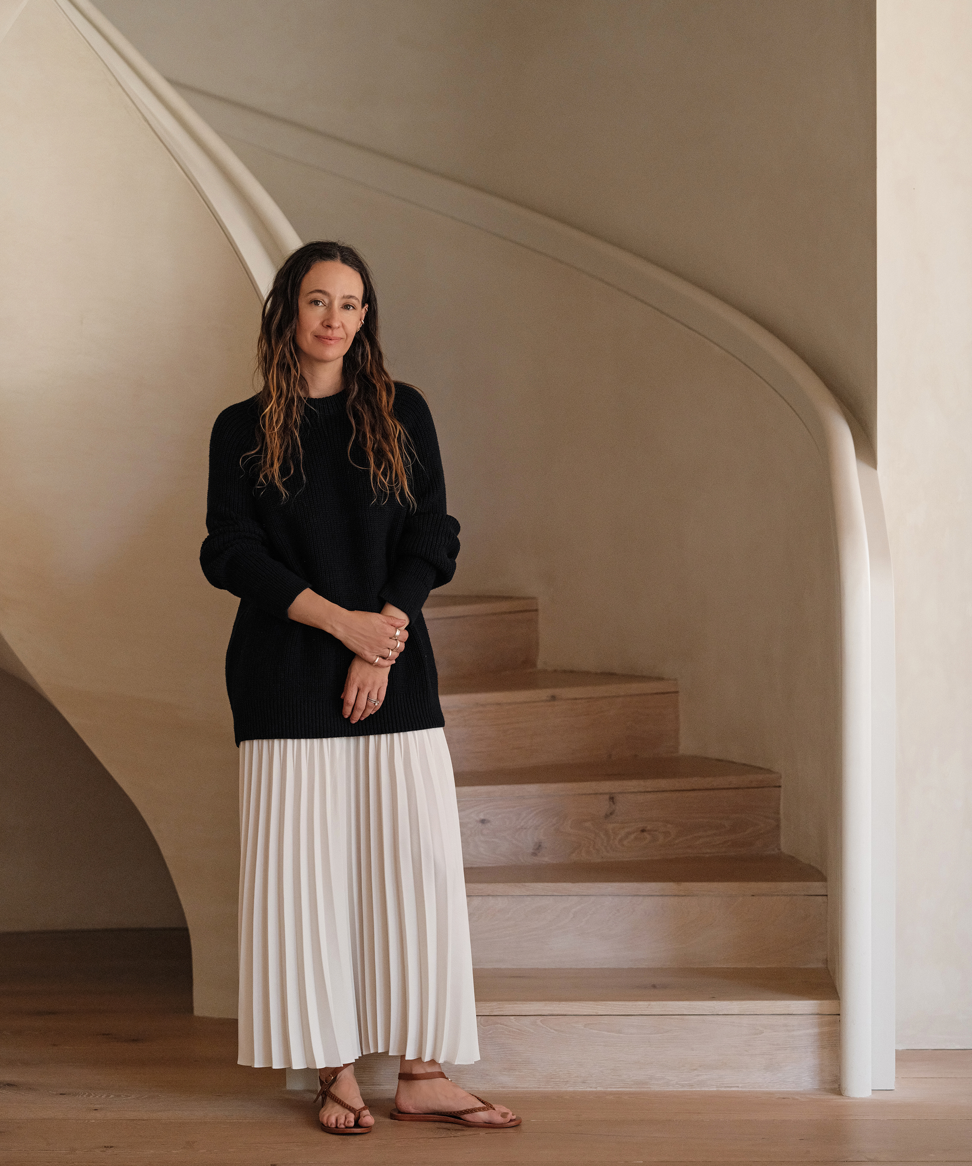 jenni leaning against a stairwell wearing a black oversized cotton fisherman sweater and an ivory pleated skirt