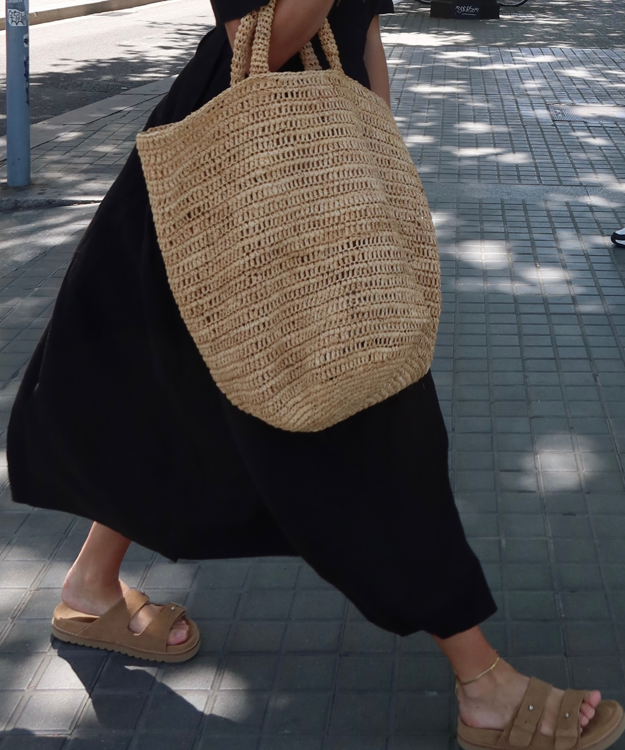 A person wearing a long black dress and tan sandals walks on a paved sidewalk, carrying a large woven straw tote bag by Lilly Sisto. Dappled sunlight and shadows are visible on the ground.