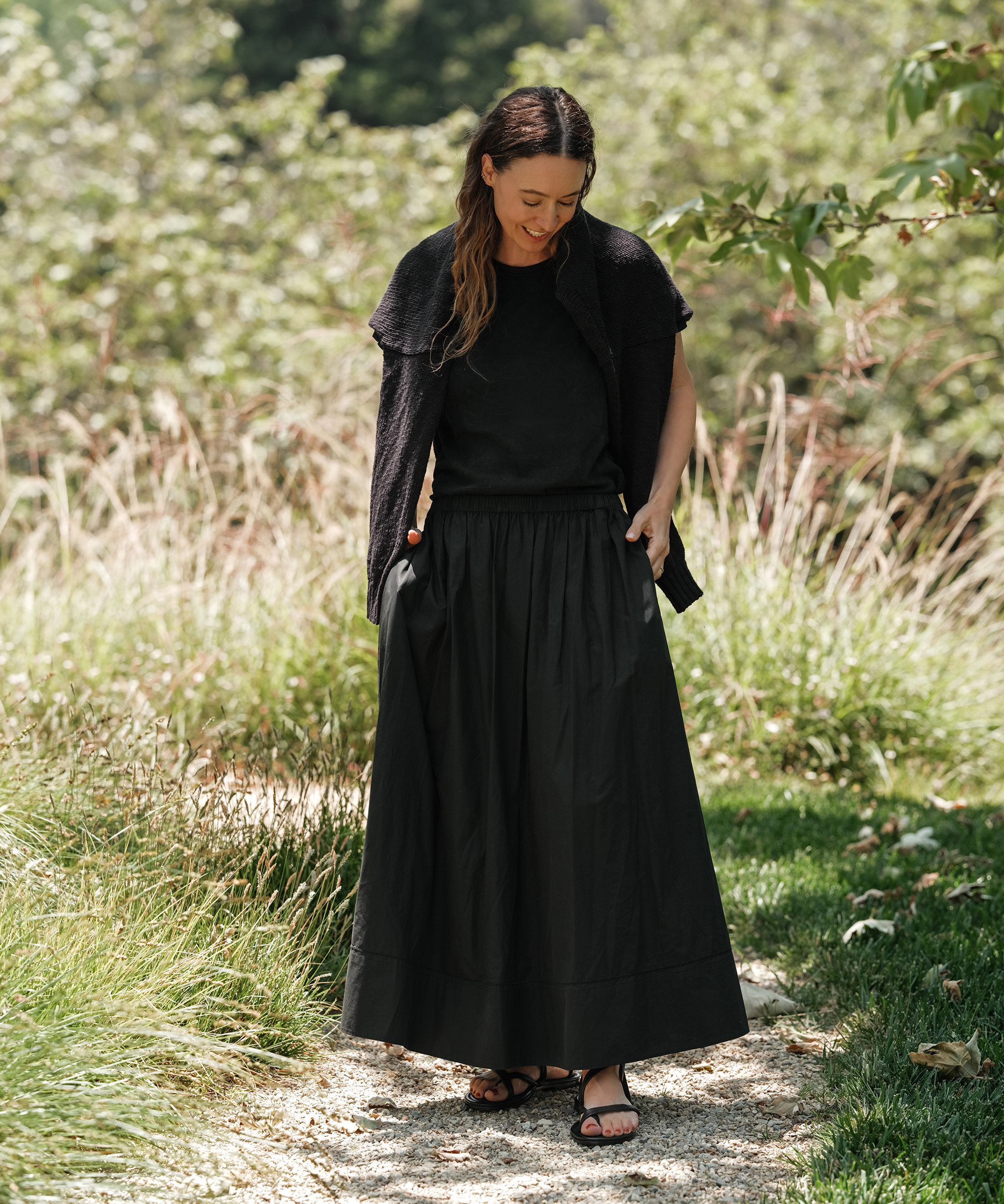 A woman stands on a garden path, looking down and smiling. She wears a black short-sleeve top, a cardigan over her shoulders, a long skirt, and sandals—an effortlessly chic choice for summer outfits. Lush greenery and tall grasses surround her.