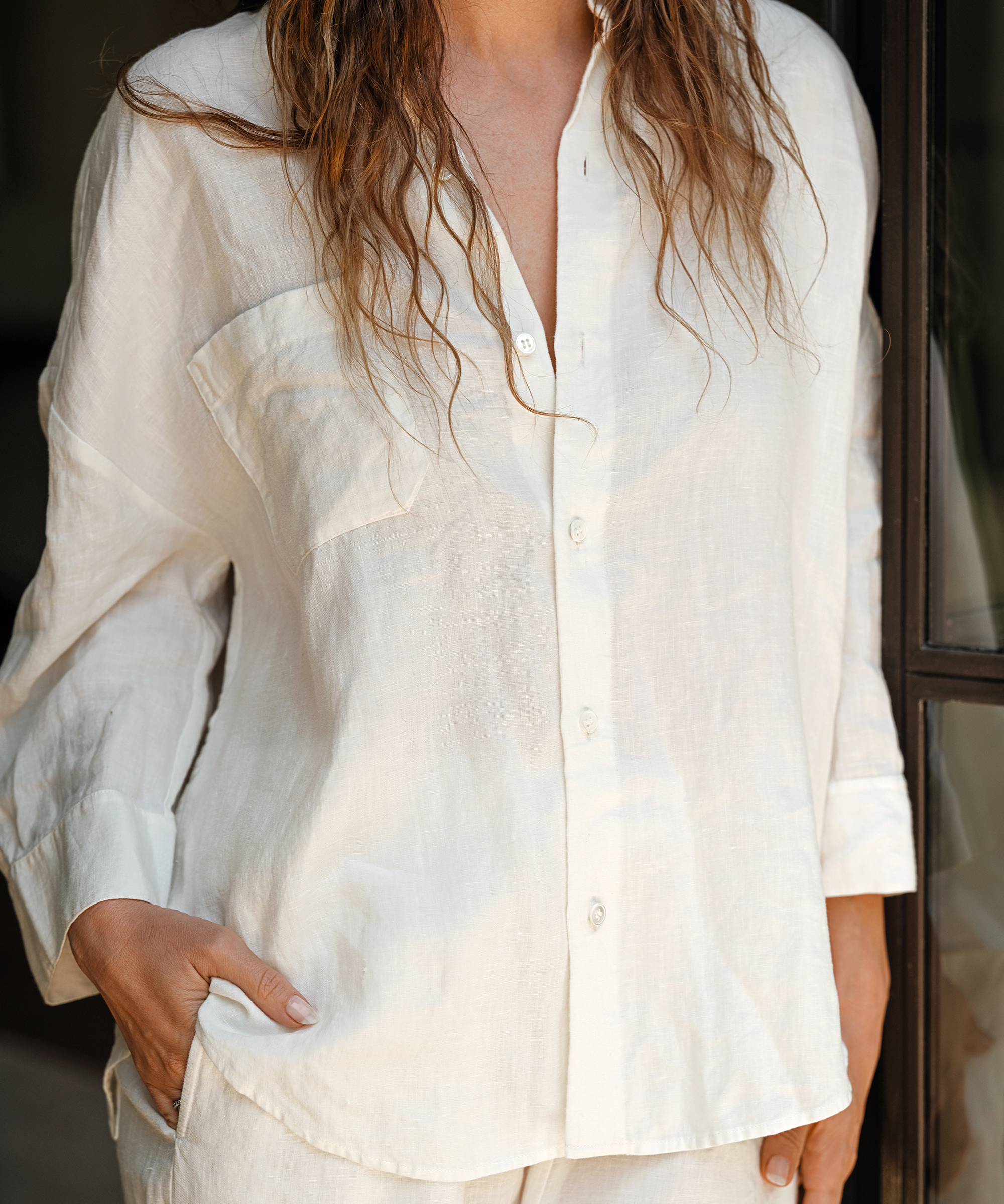 A person with wavy hair wearing a loose, off-white button-up shirt and white pants stands indoors with one hand in their pocket, partially facing the camera—a relaxed look perfect for summer outfits. Their face is not visible.