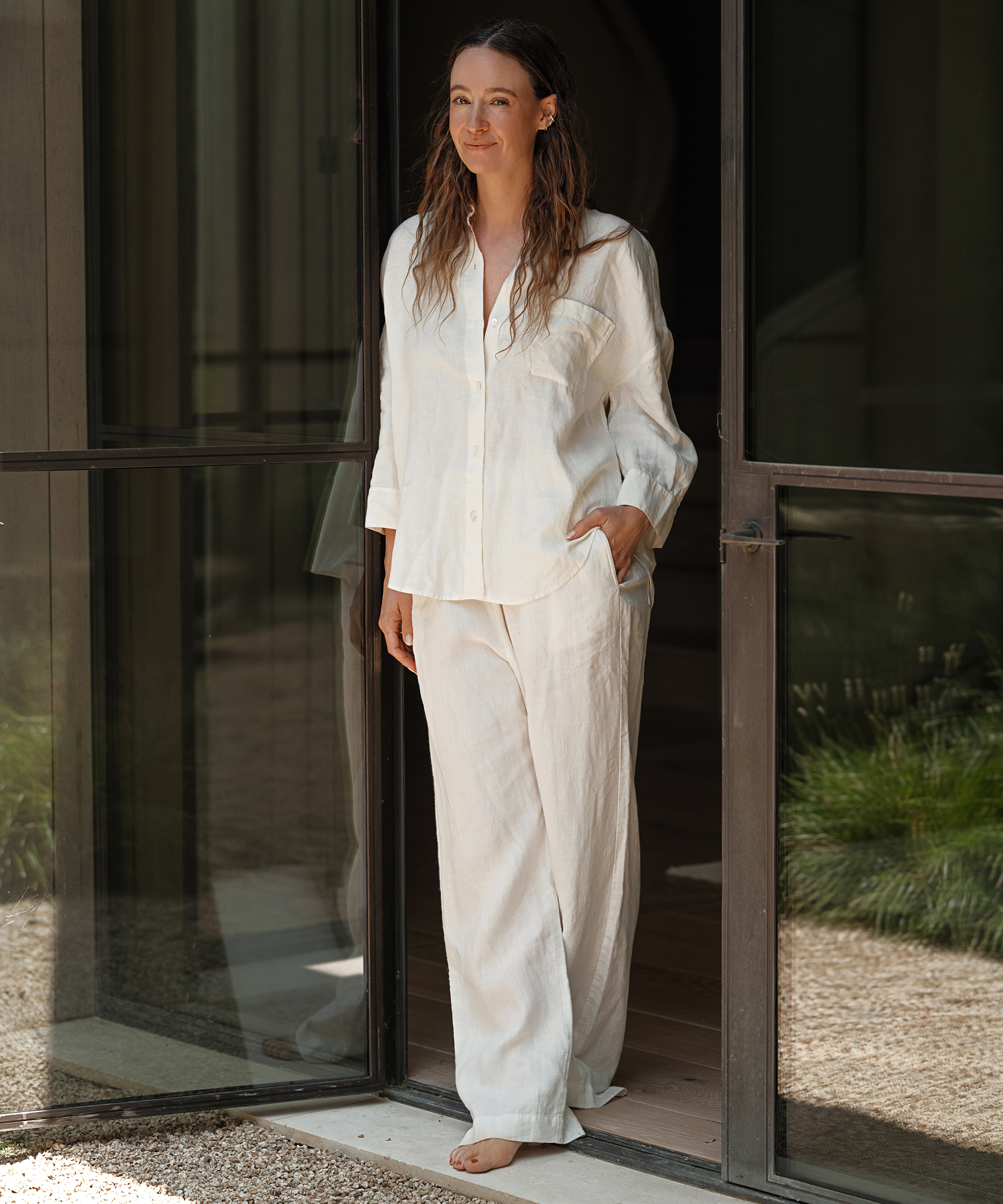 A woman wearing a loose, white linen shirt and pants stands barefoot in a doorway, smiling slightly. Sunlight filters in, greenery is visible outside—capturing the relaxed elegance of effortless summer outfits.