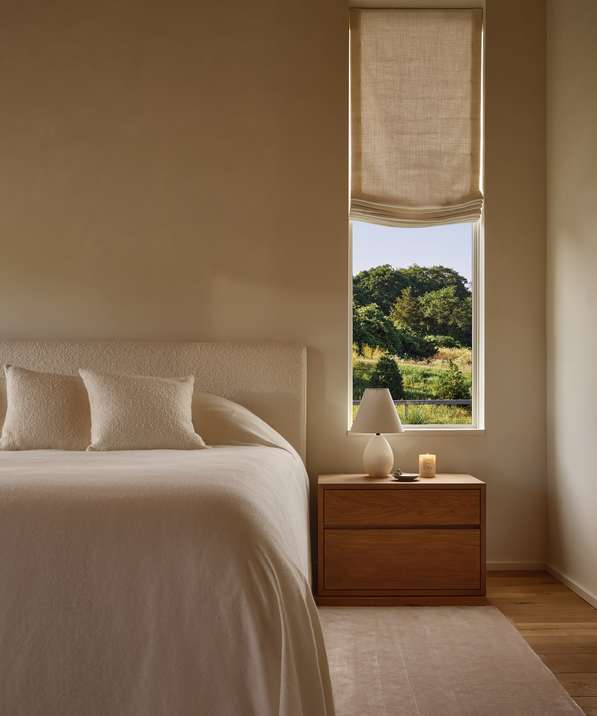 Minimalist bedroom featuring a bed with white bedding and pillows, a wooden nightstand topped with a white lamp and candle, and a window with a partially raised beige shade revealing green trees outside.