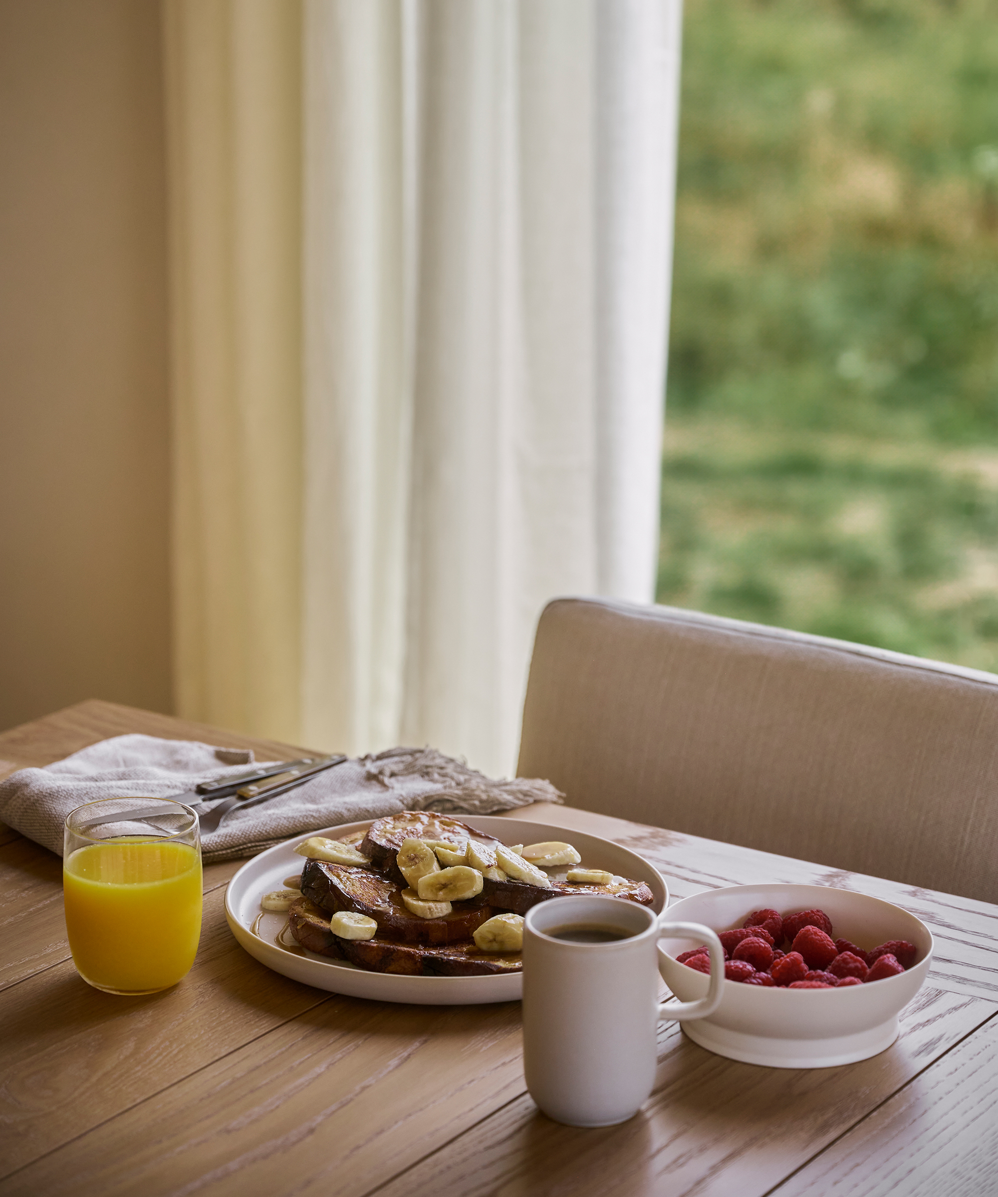 A breakfast scene on a wooden table with French toast topped with banana slices, a bowl of raspberries, and fresh orange juice showcases the charm of farm to table recipes by a window with sheer curtains and lush greenery outside.