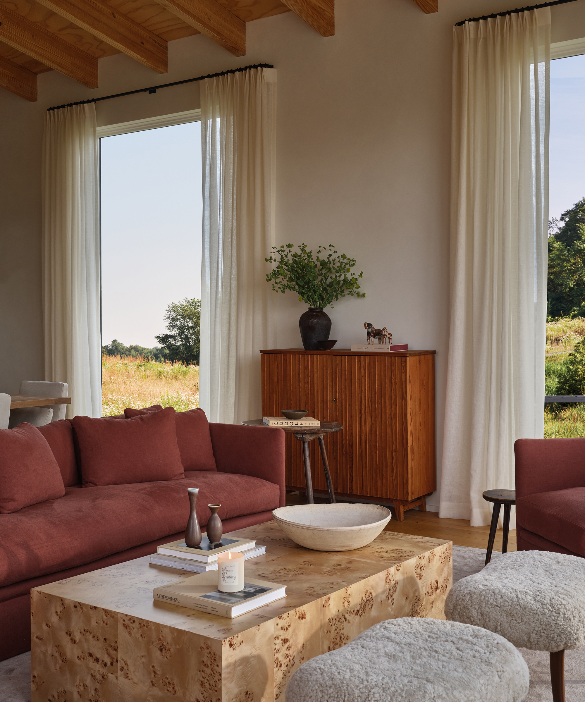 A cozy living room with a rust-colored sofa, wooden coffee table with books and decor, two textured stools, and a wooden cabinet with a plant evokes Jenni Kayne farmhouse style. Large windows with white curtains overlook a green landscape.