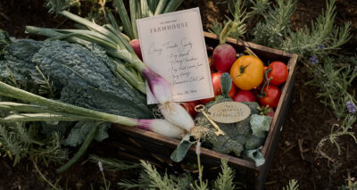 A wooden crate filled with fresh vegetables—kale, green onions, radishes, tomatoes, and broccoli—sits on the ground. A handwritten farm to table recipe, a gold key, and a metal tag reading “The JeniKayke Farmhouse” rest on top.