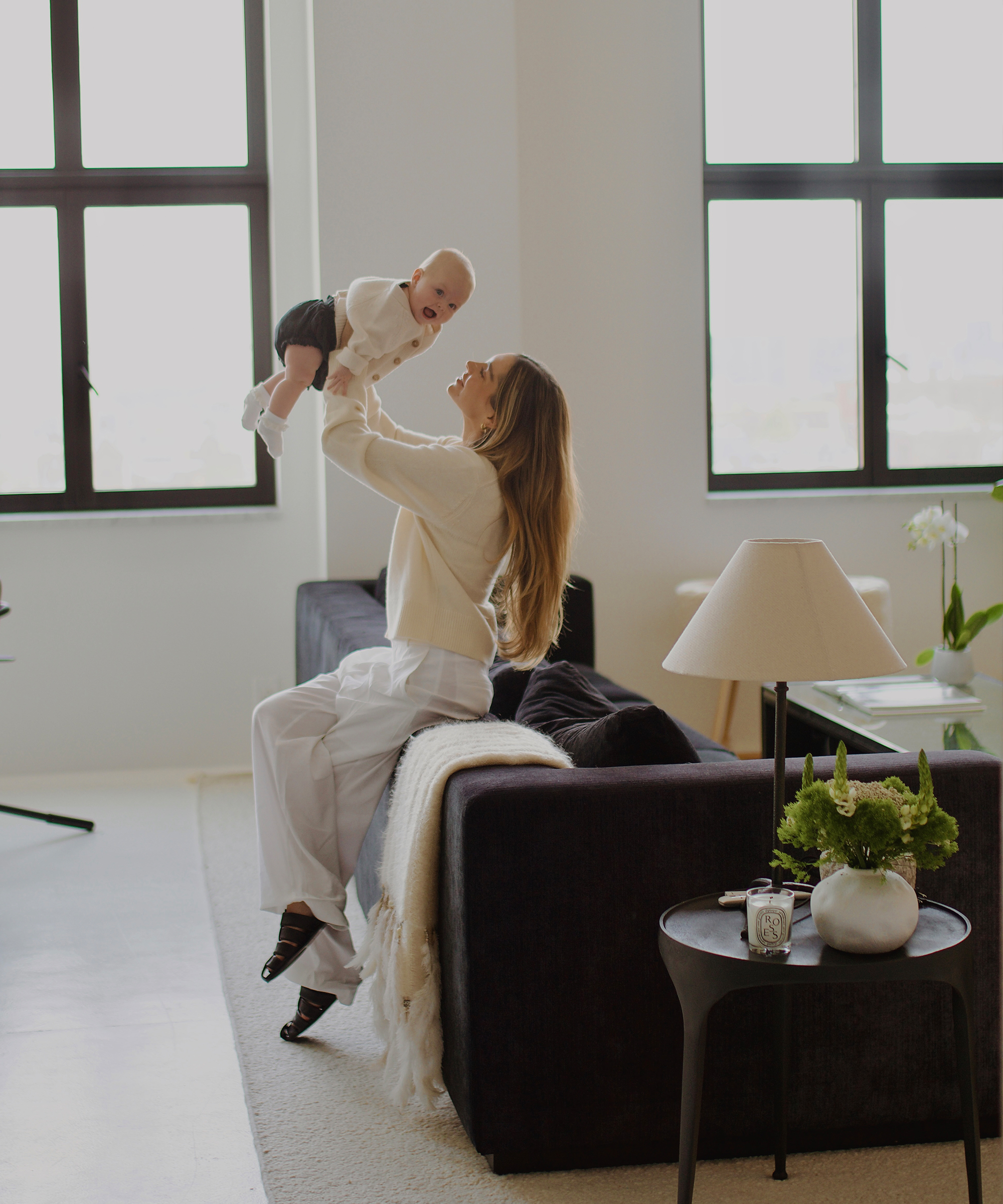 lauren burke wearing white trousers and an ivory cashmere cardigan holding her baby in her arms sitting on a charcoal couch in her living room